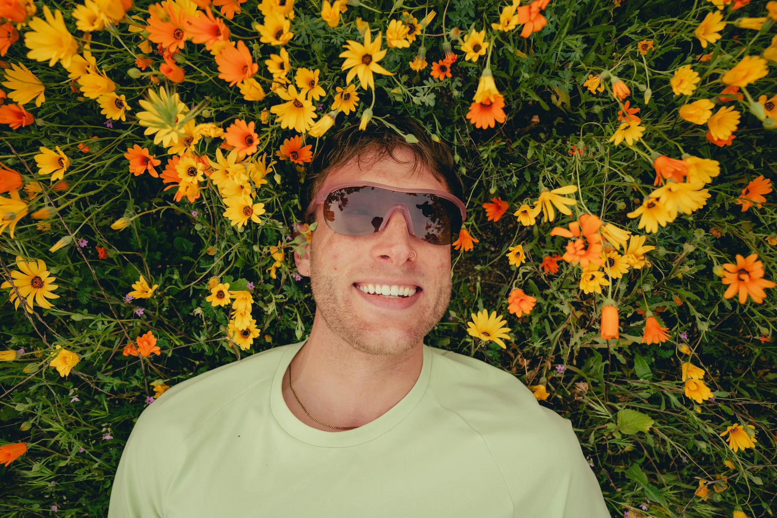 A smiling man lying on a bed of yellow and orange flowers, wearing sunglasses and a light-colored shirt.
