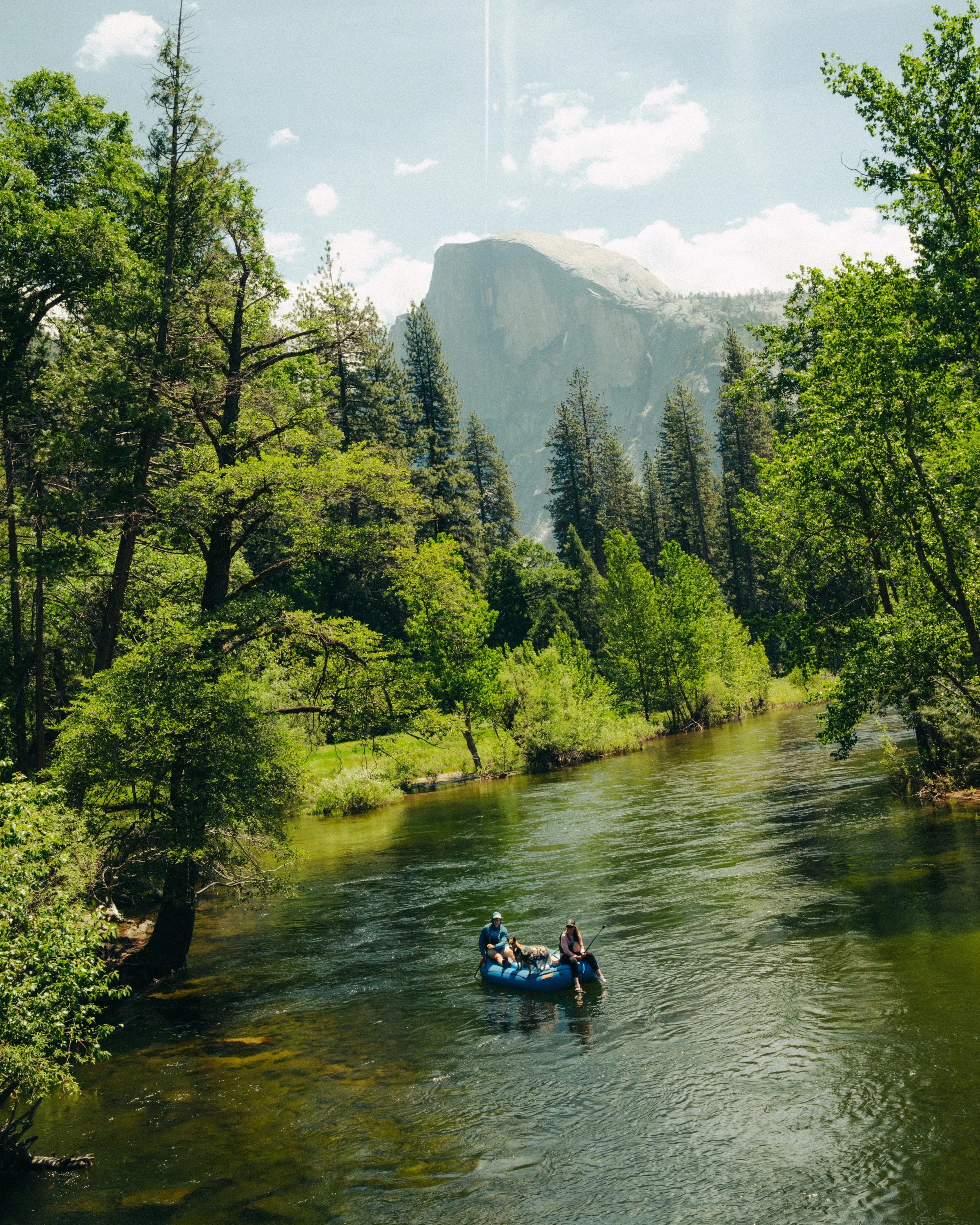 People wearing life jackets on a blue inflatable boat floating on a river surrounded by green trees, with mountains and a partly cloudy sky in the background.