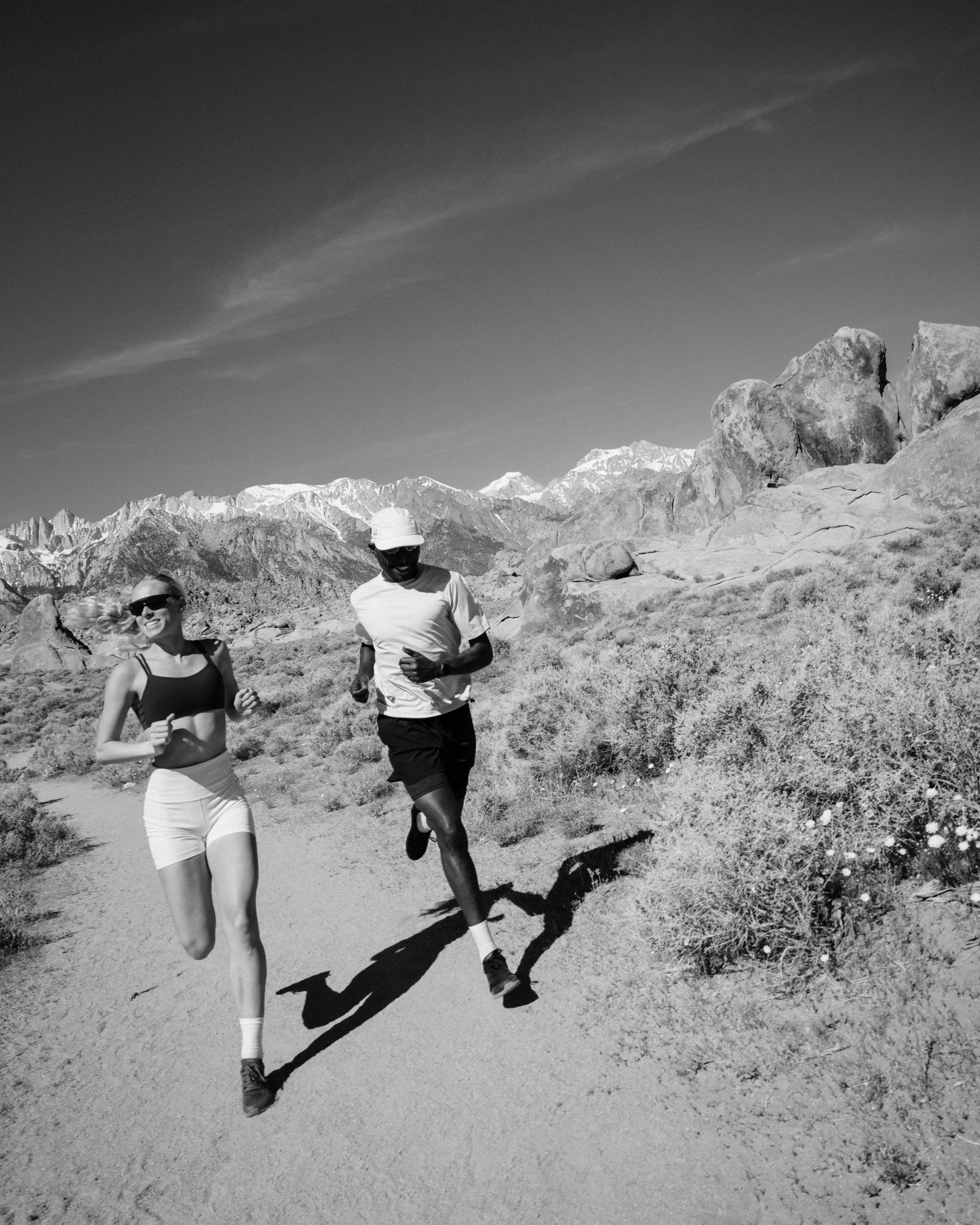 Two people jogging on a mountain trail with rocky terrain, large boulders, and snow-capped peaks in the distance under a clear sky.
