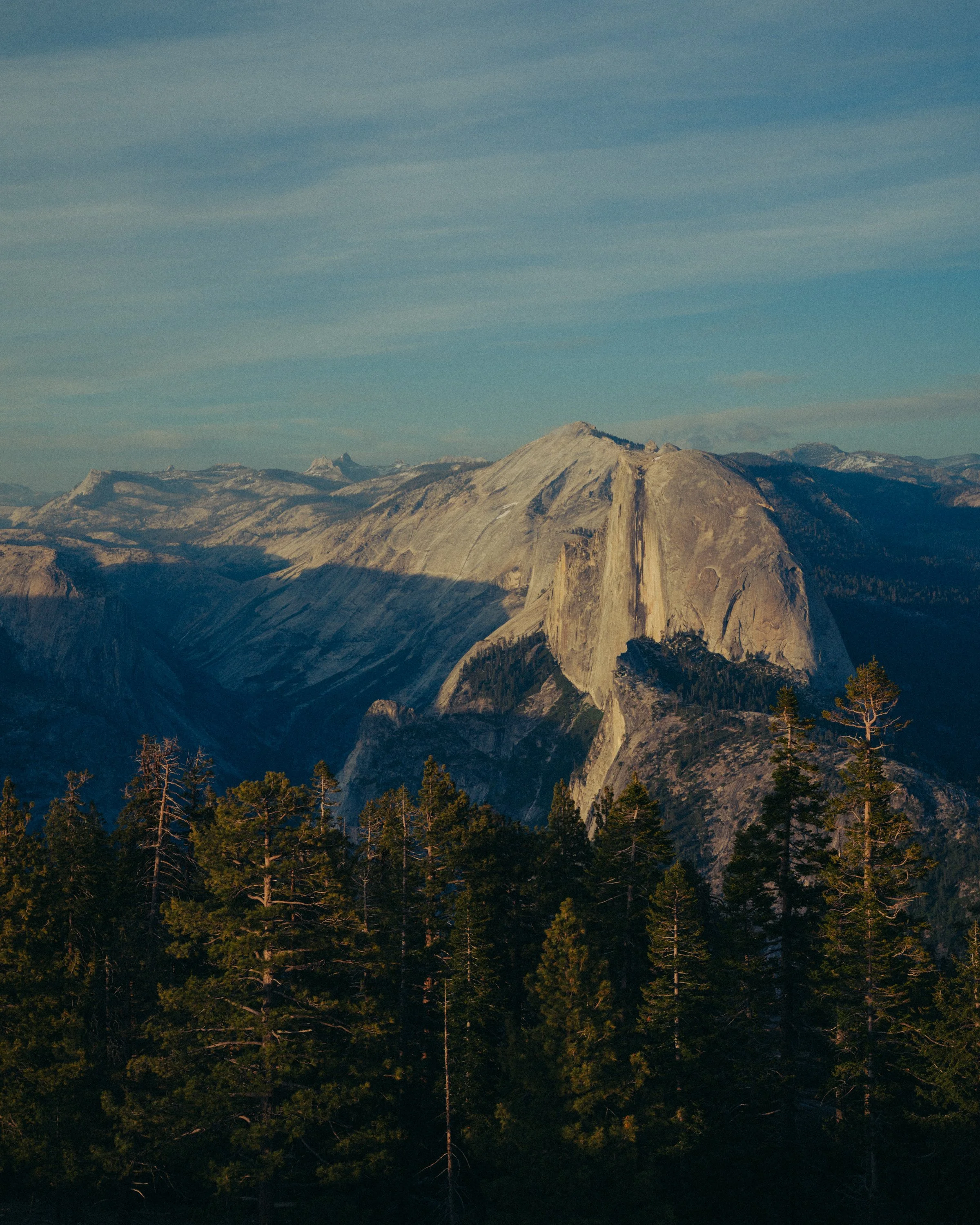 A mountain landscape with large granite cliffs, rolling hills, and a forest of evergreen trees in the foreground under a partly cloudy sky.