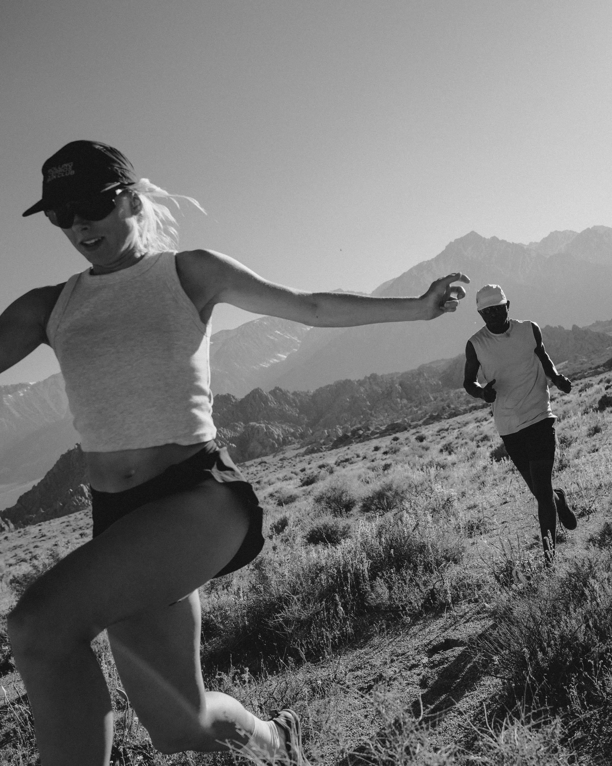A black and white photo of two people running outdoors in a mountainous area with dry grass and shrubs.