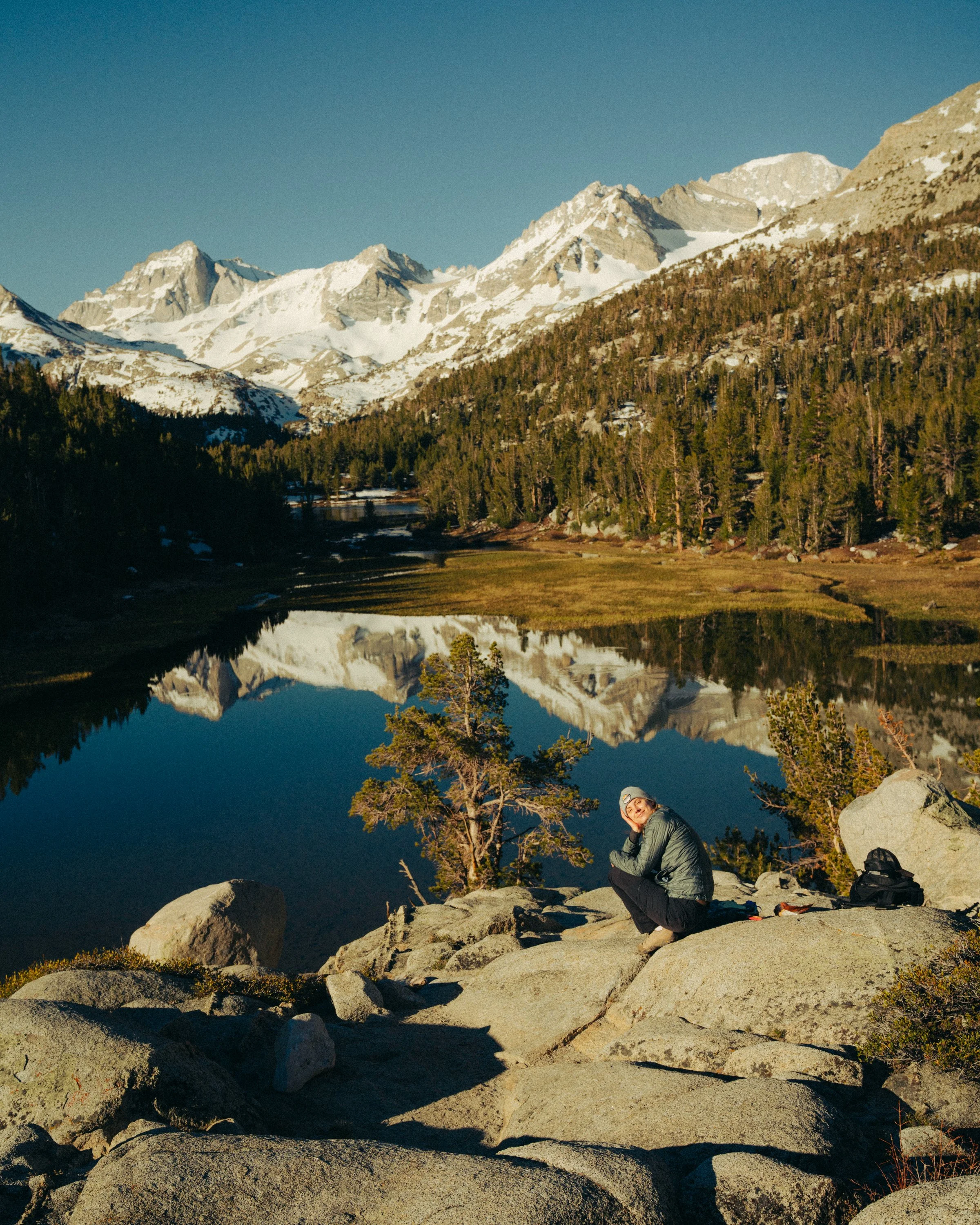 A person sitting on rocks near a calm lake surrounded by tall pine trees and snow-capped mountains under a clear blue sky.