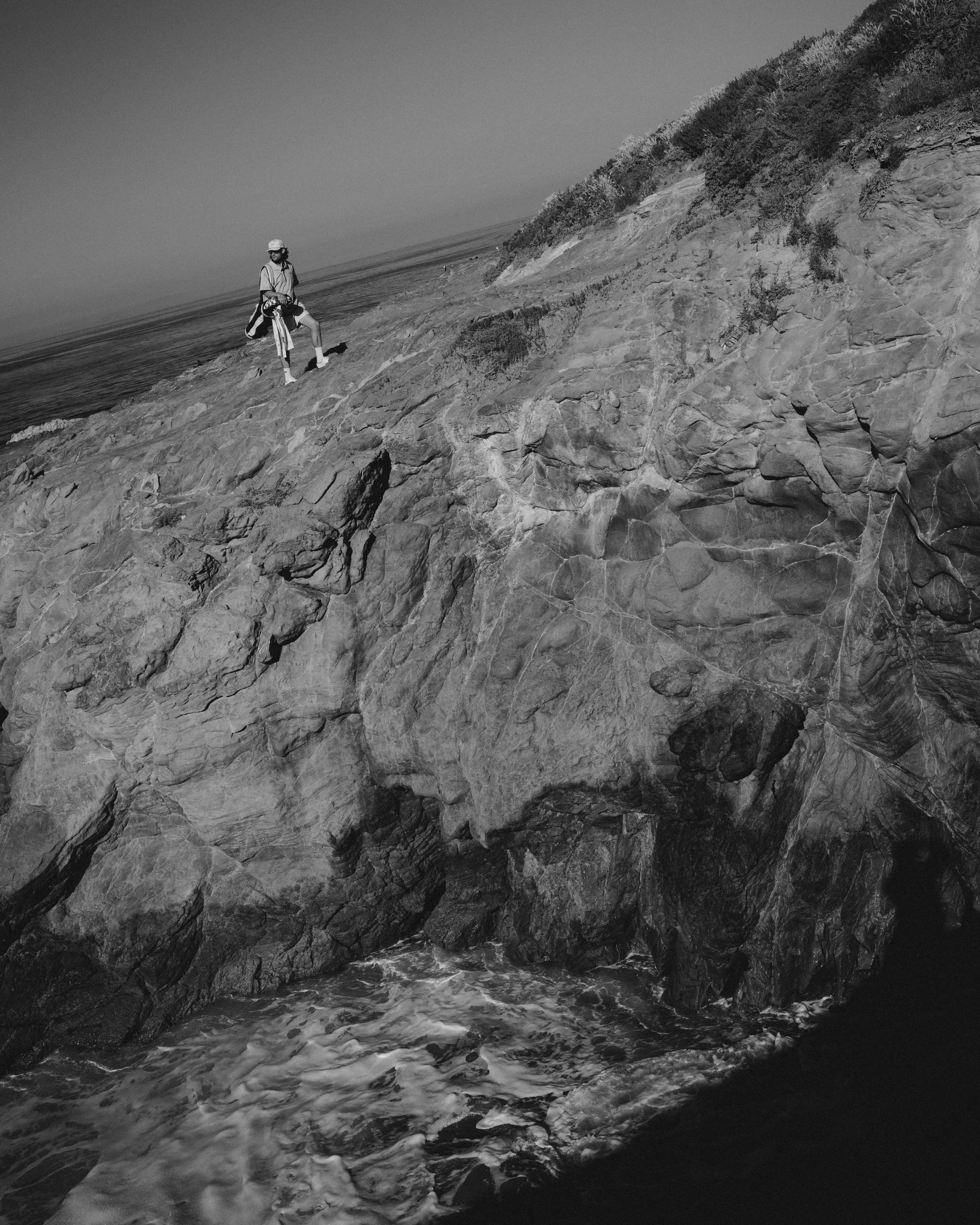 A person standing on a rocky cliff near the ocean, dressed in outdoor attire and wearing a helmet, with waves crashing below.