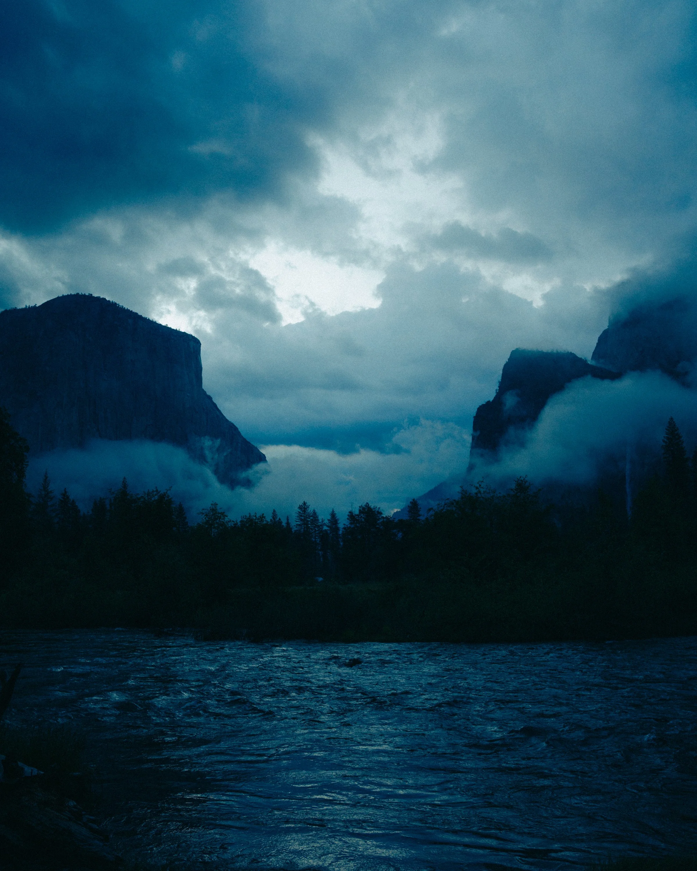 A view of cloudy blue sky over mountains with fog and a body of water in the foreground.