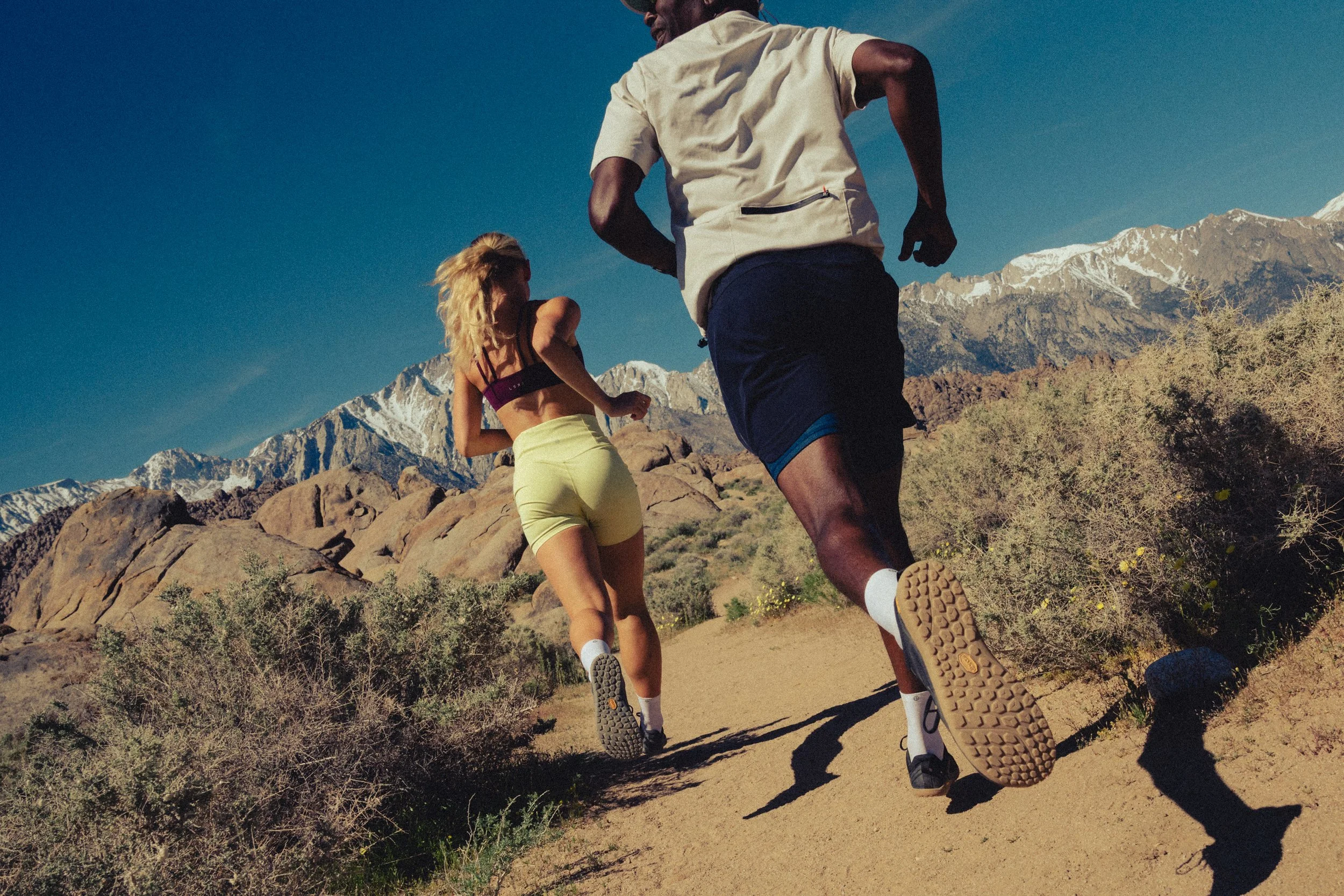 Two runners, a man and a woman, running on a trail in a mountainous desert landscape with snow-capped peaks in the background. The man is wearing a light-colored shirt and dark shorts, and the woman has blonde hair and is wearing a sports bra and yellow shorts.