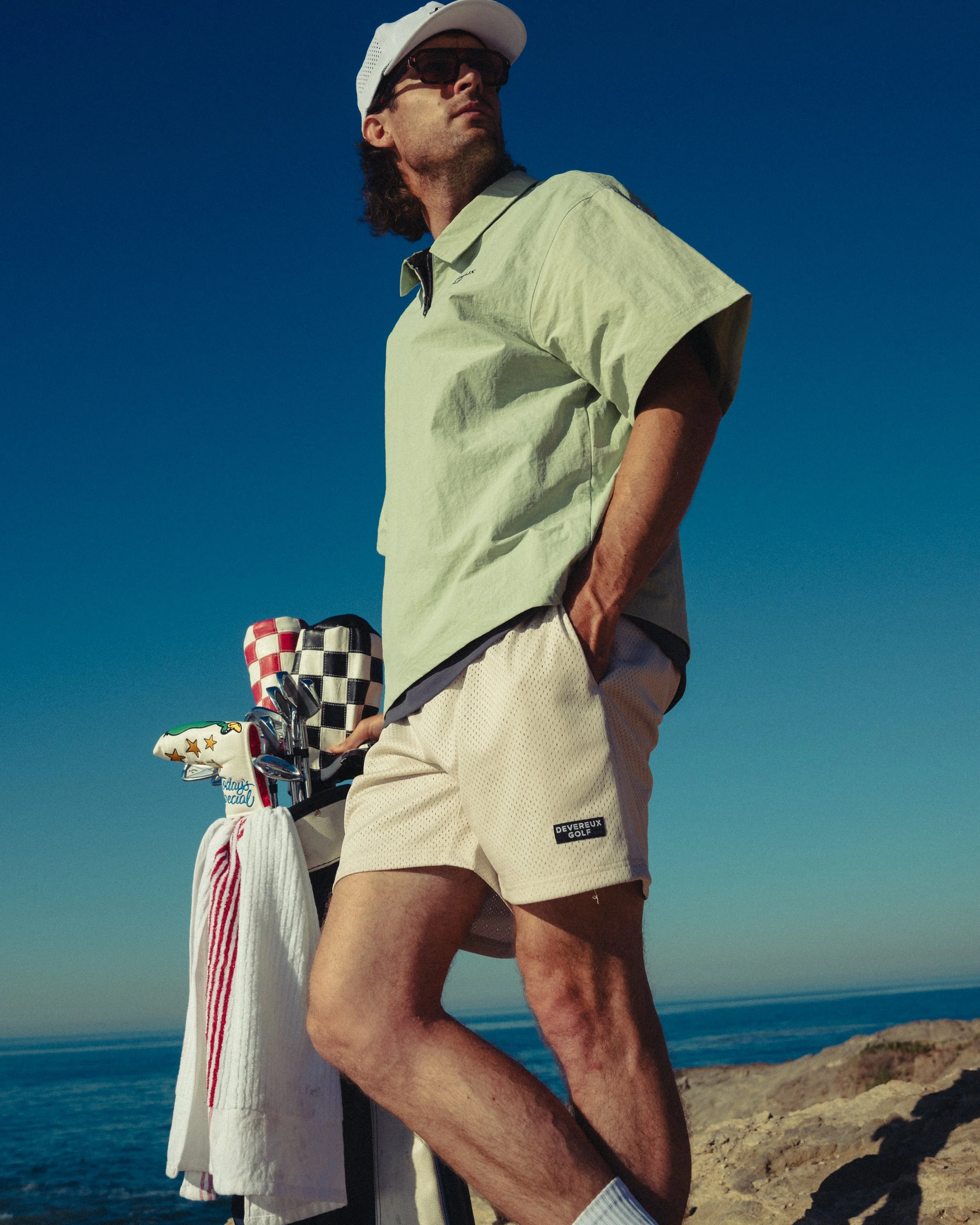 A man wearing a white cap, sunglasses, and a light green shirt stands on a rocky area near the sea. He has a set of golf clubs with a towel hanging from them, and the sky is clear and blue.