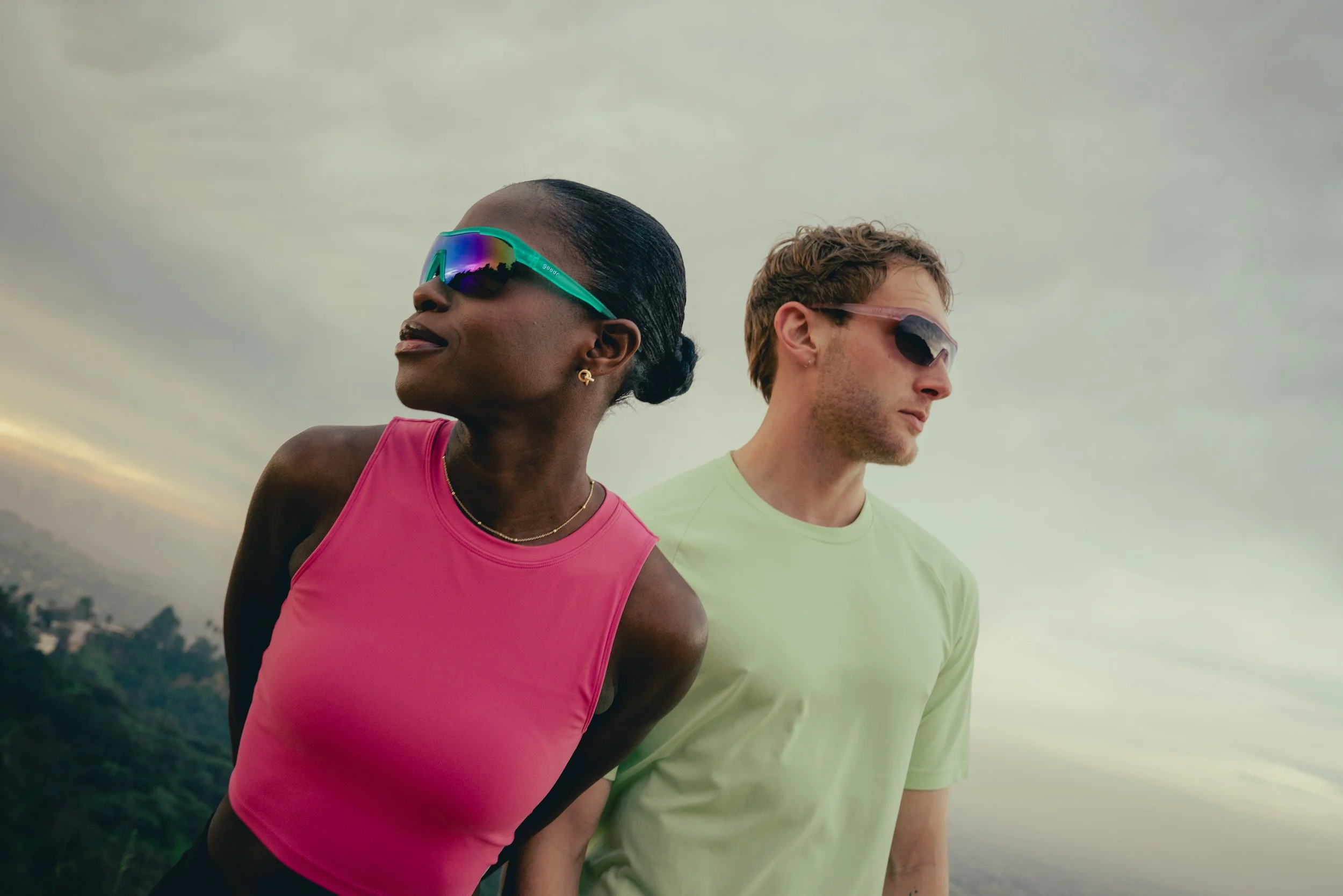 A woman in a pink athletic top and man in a light green t-shirt standing outdoors with a cloudy sky background.