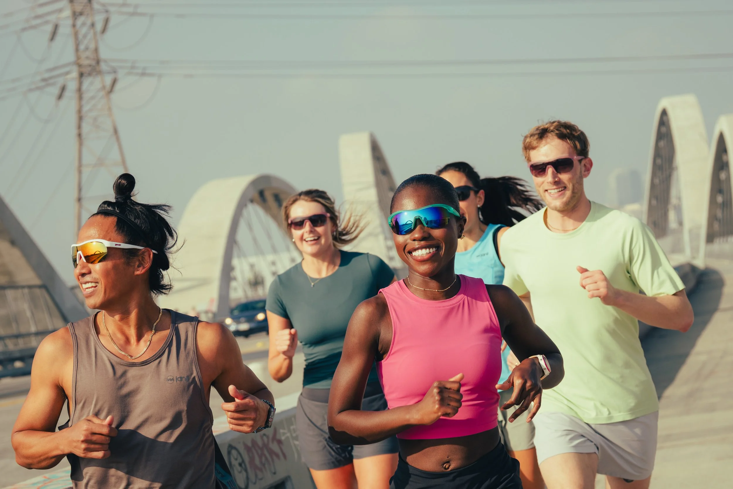 Group of diverse runners jogging outdoors on a bridge with modern arches, wearing athletic clothing and sunglasses, under a clear sky.