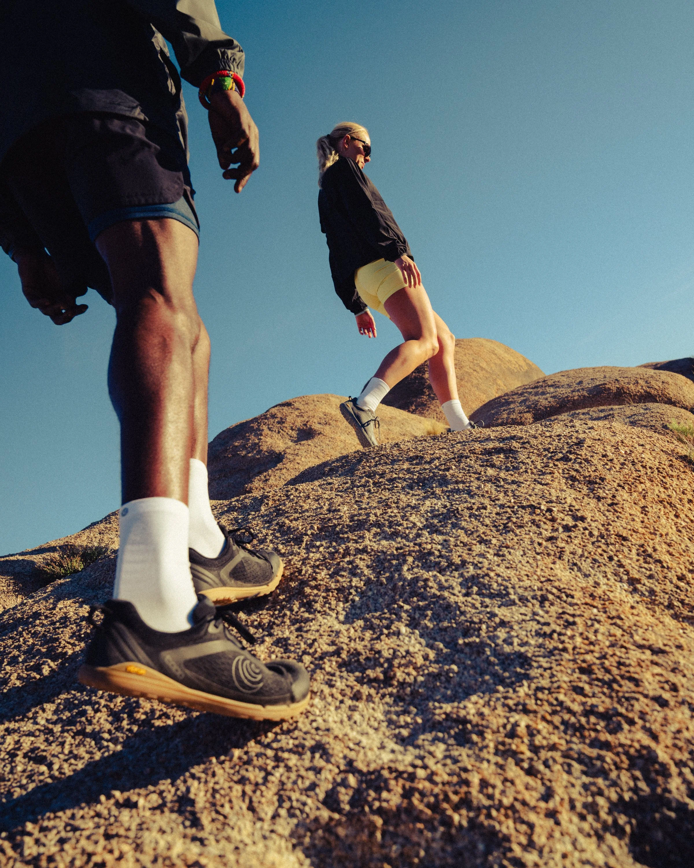 A person climbing rocky terrain wearing athletic shoes, yellow shorts, and a black jacket, while another person is standing nearby, dressed in dark shorts and a jacket, against a clear blue sky.