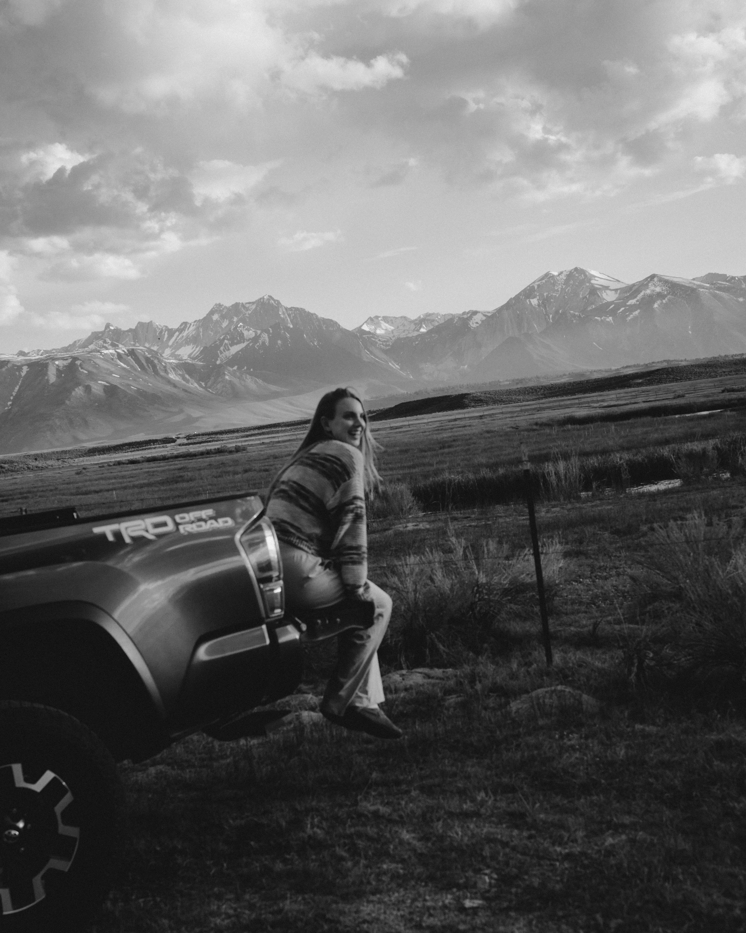 A woman sitting on the side of a road beside a vehicle, with mountains and clouds in the background. The woman is smiling and looking off into the distance, dressed casually, in black and white photo.