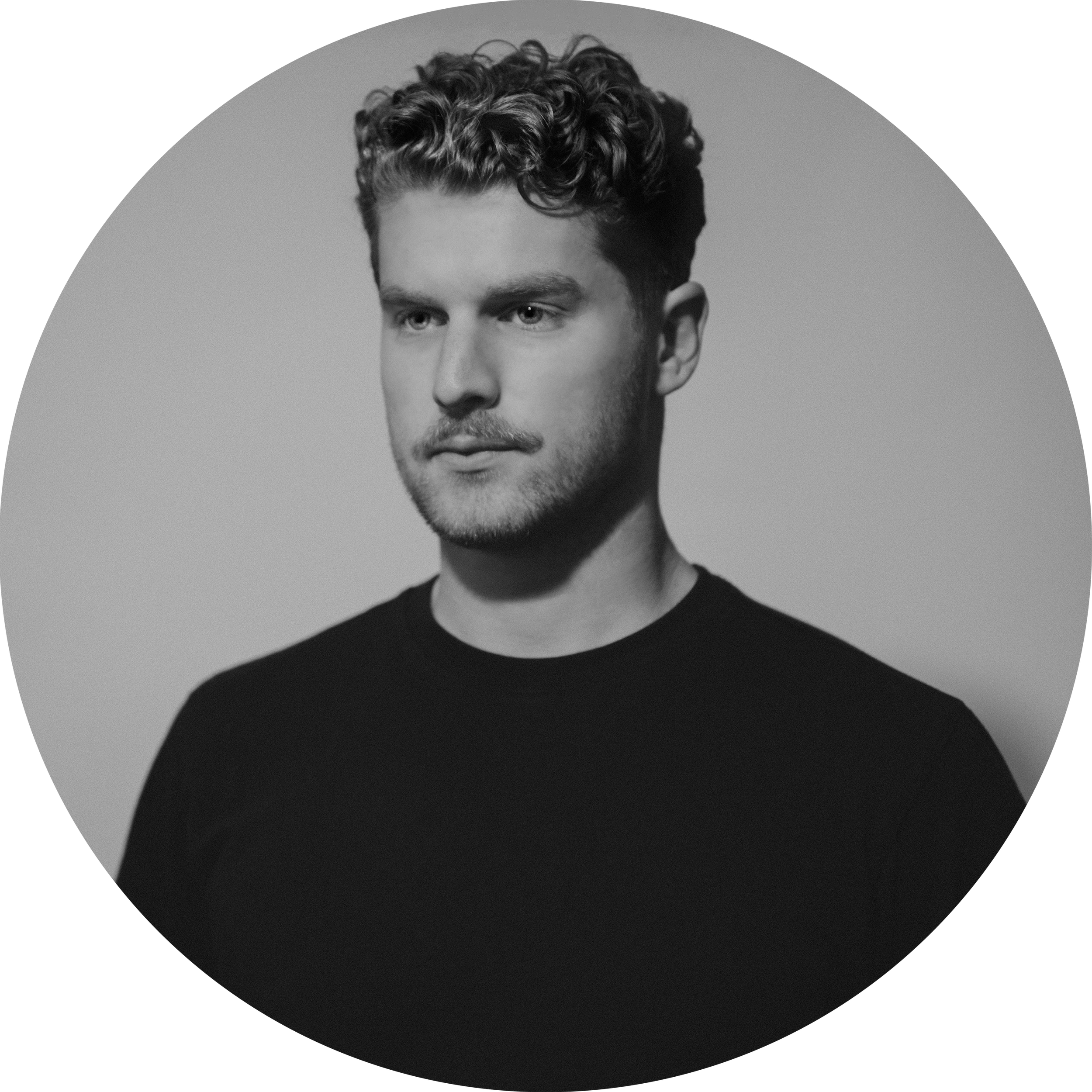 A young man with short, curly hair and a light beard, wearing a black shirt, looking slightly to the side in a black-and-white portrait.