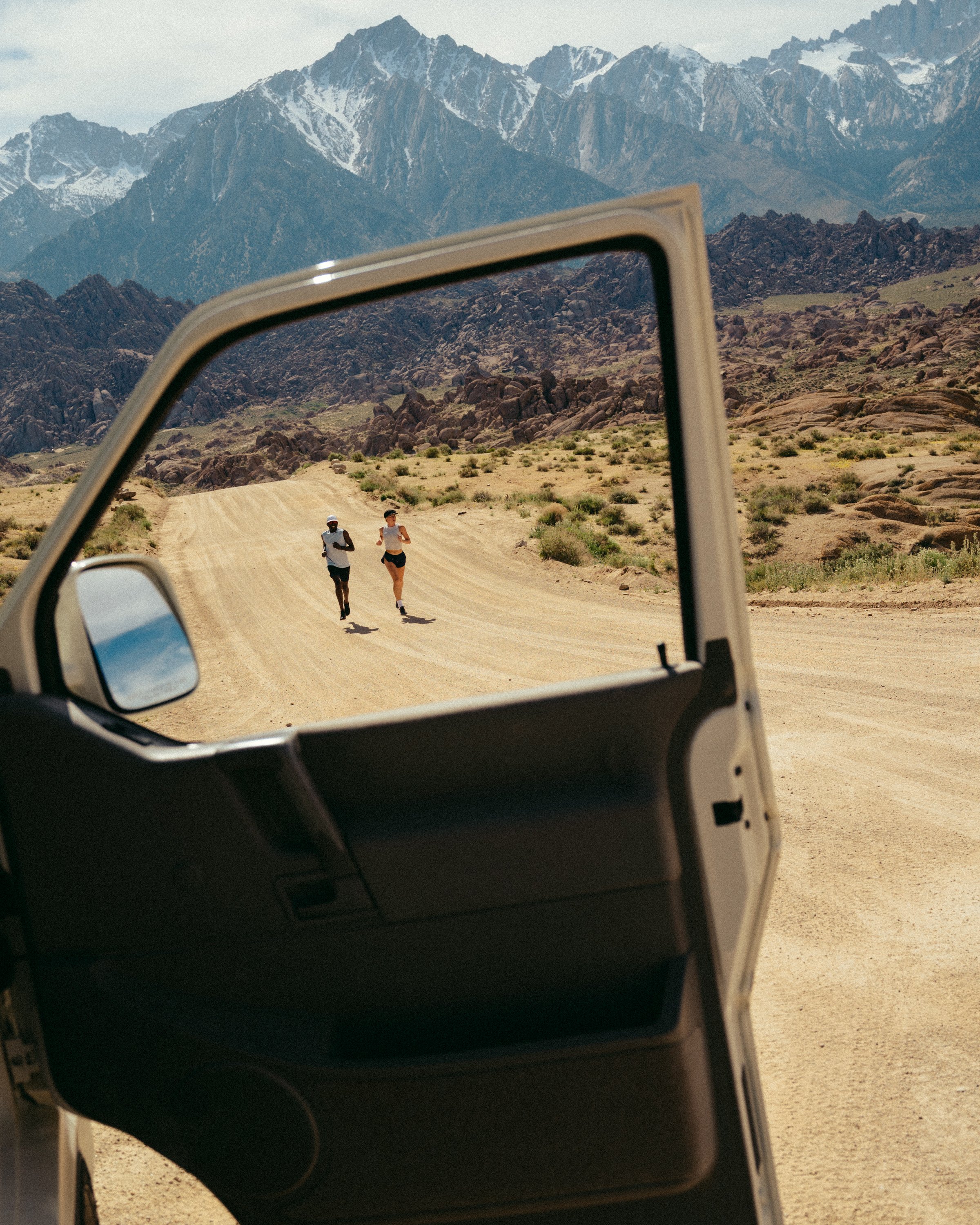 A desert landscape with rocky terrain, two runners on a dirt path, and snow-capped mountains in the background, viewed through the side mirror of a vehicle.