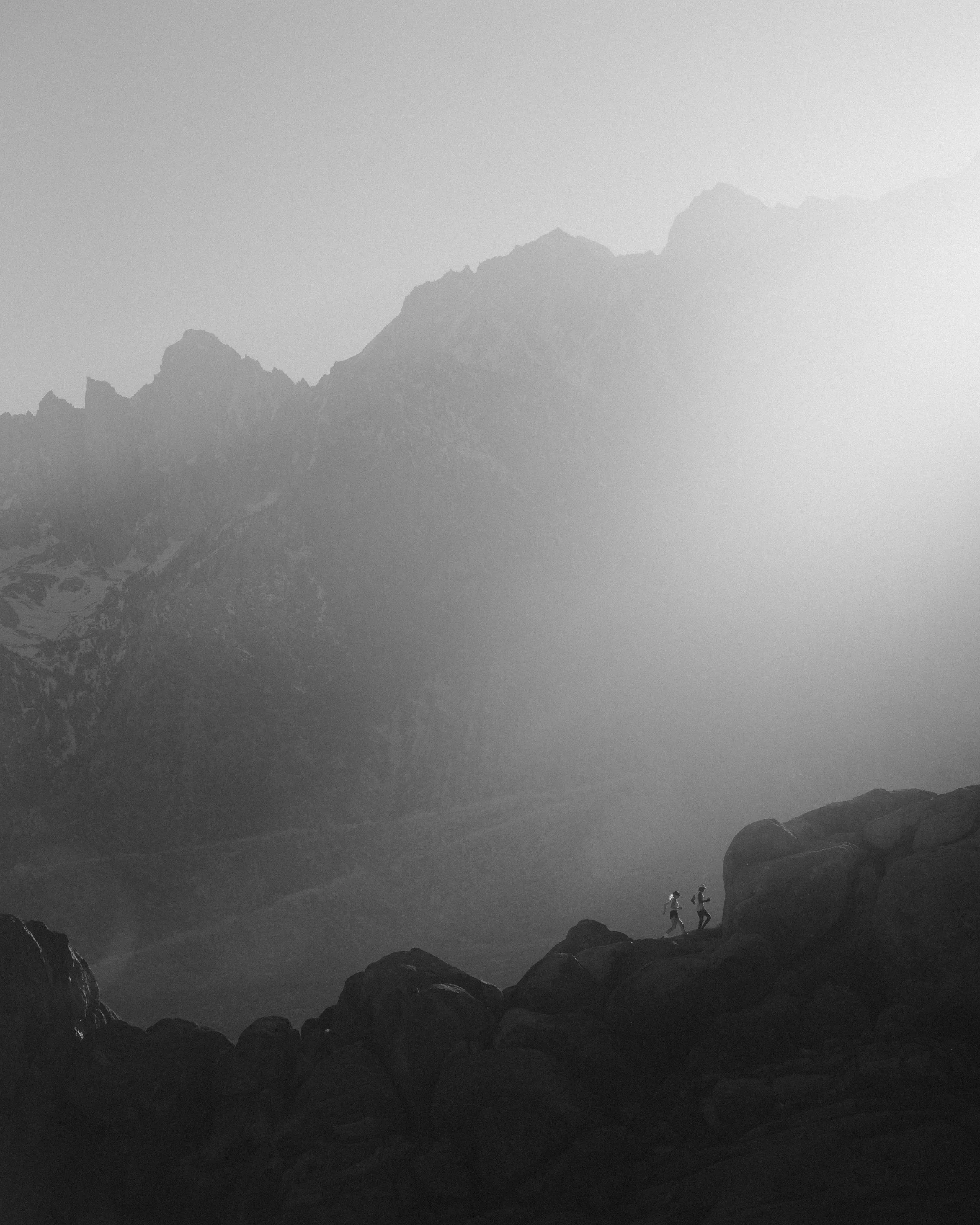 Black and white photograph of two hikers walking on a rocky terrain in a mountainous landscape with large rocks and peaks in the distance.