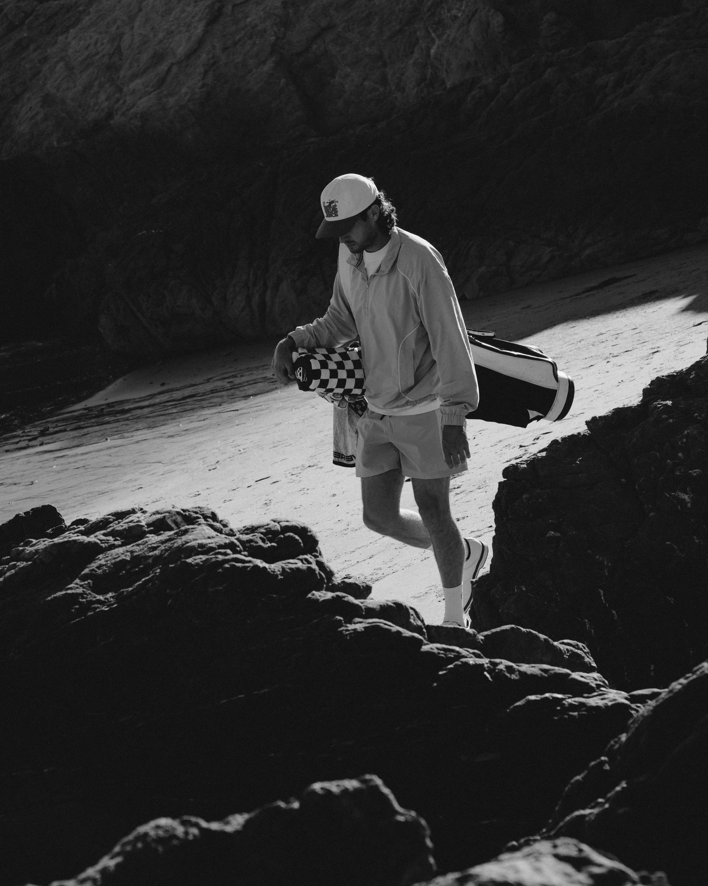 A man walks on rocky terrain at the beach, carrying a golf club and golf bag, wearing a cap, jacket, shorts, and sneakers, with rocky formations in the background.