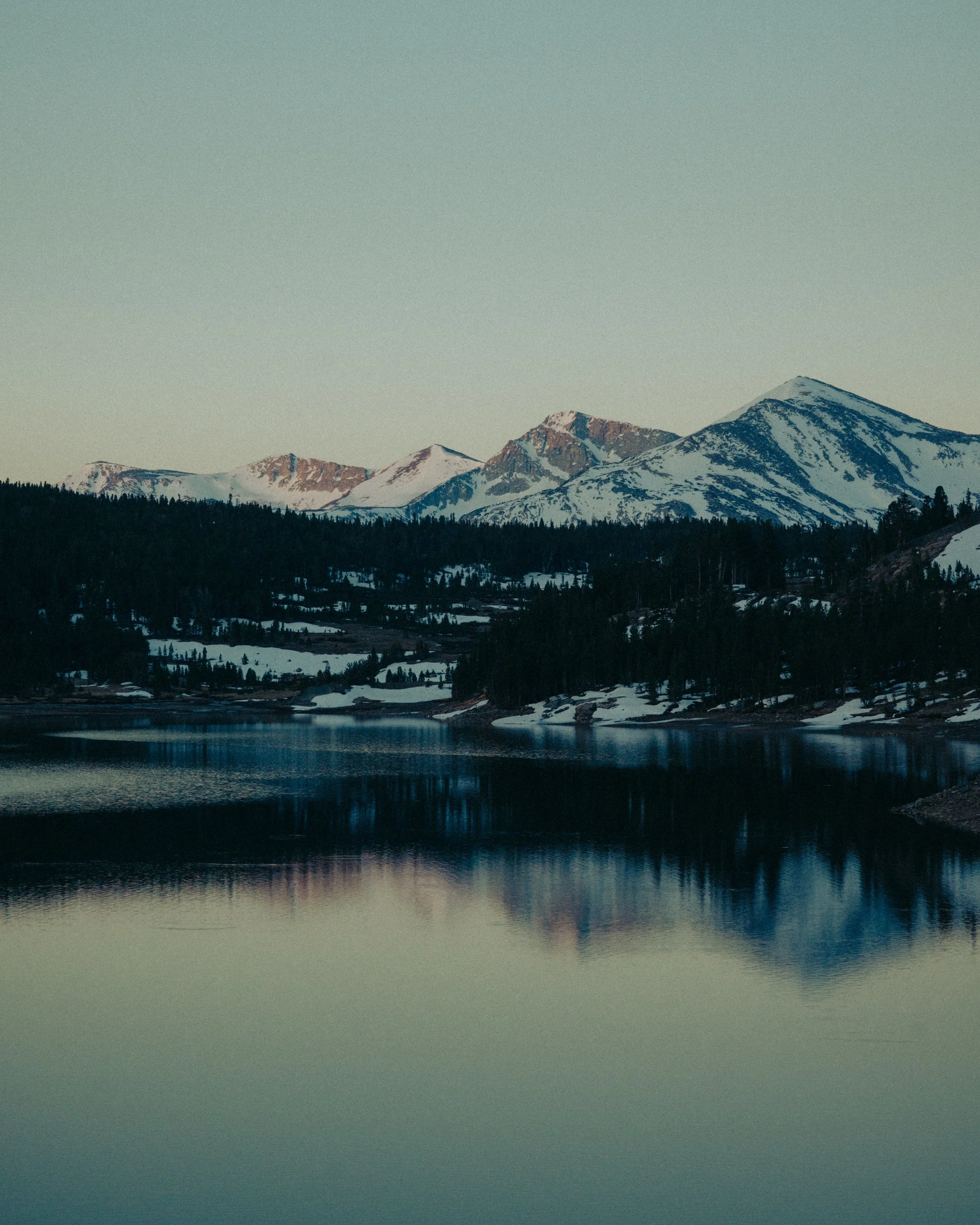 Snow-capped mountains with dark forested slopes and a body of water in the foreground under a clear sky.