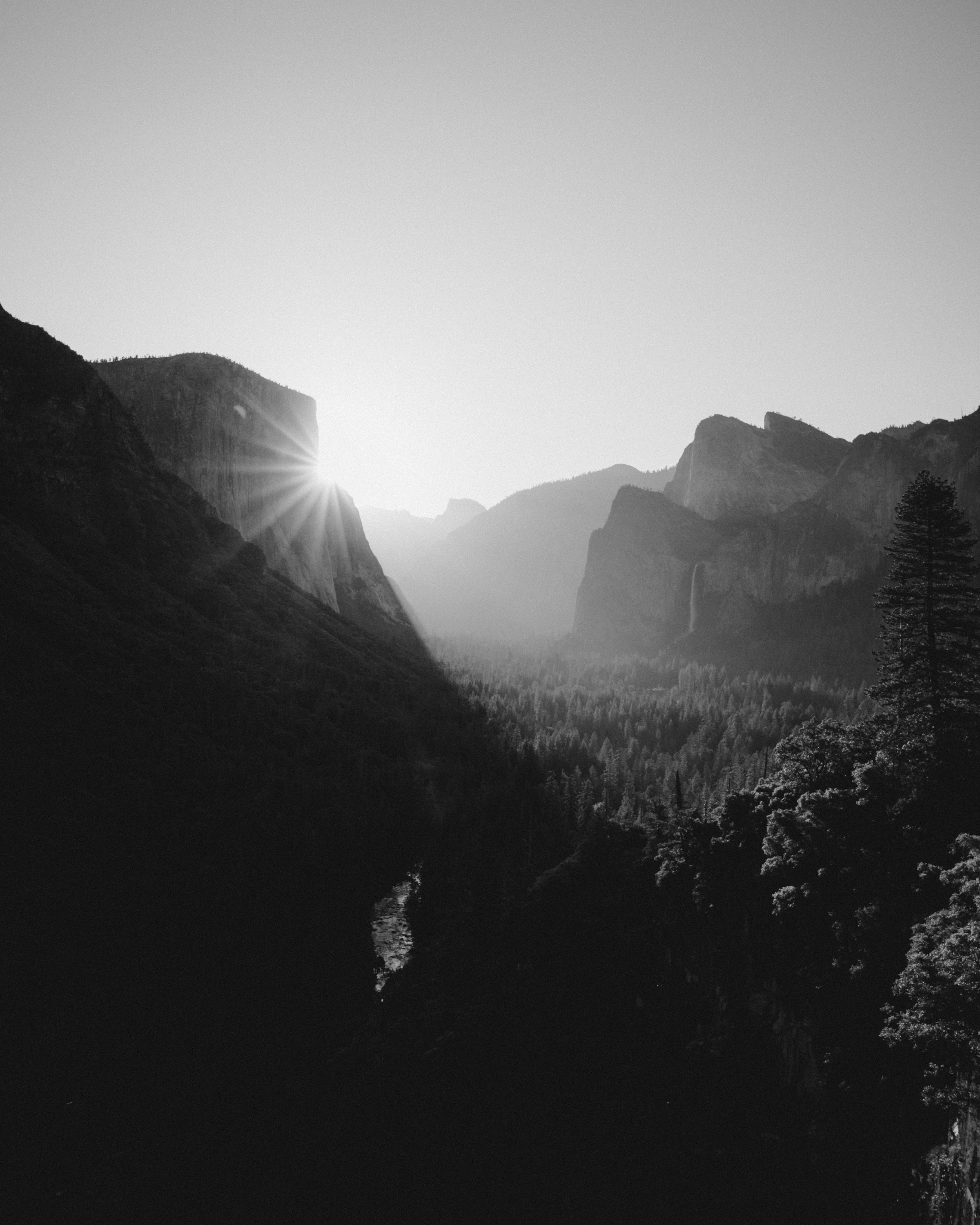 Black and white photo of a mountainous landscape with the sun setting behind the mountains, creating a glare and long shadows.