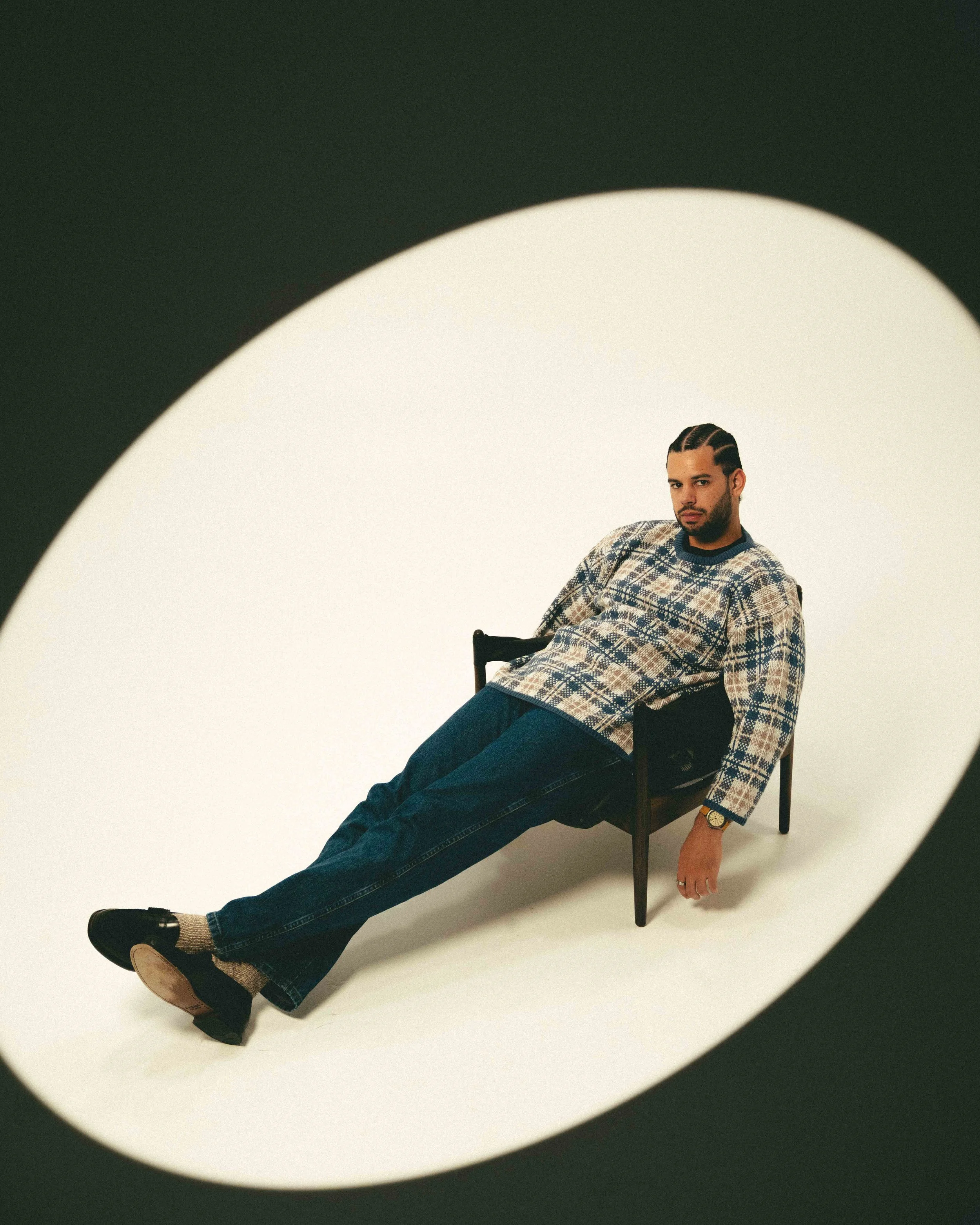 Man reclining on a small black chair, viewed through a circular opening, wearing a patterned sweater, jeans, and shoes, with braided hair and a serious expression.