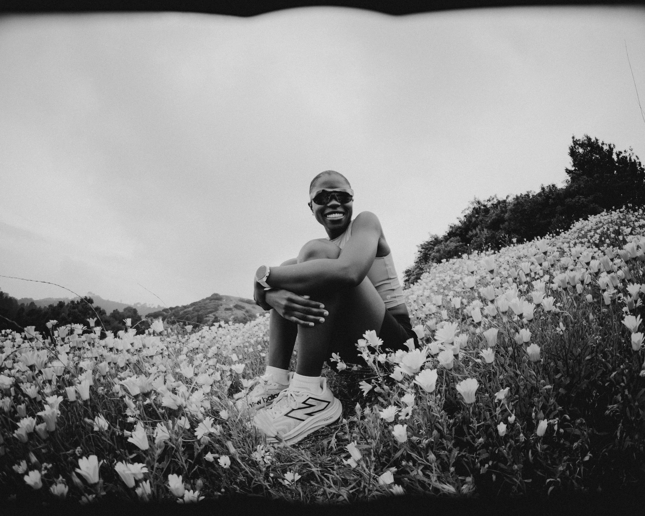 A person sitting in a field of flowers, smiling, wearing sunglasses, a tank top, shorts, and sneakers.