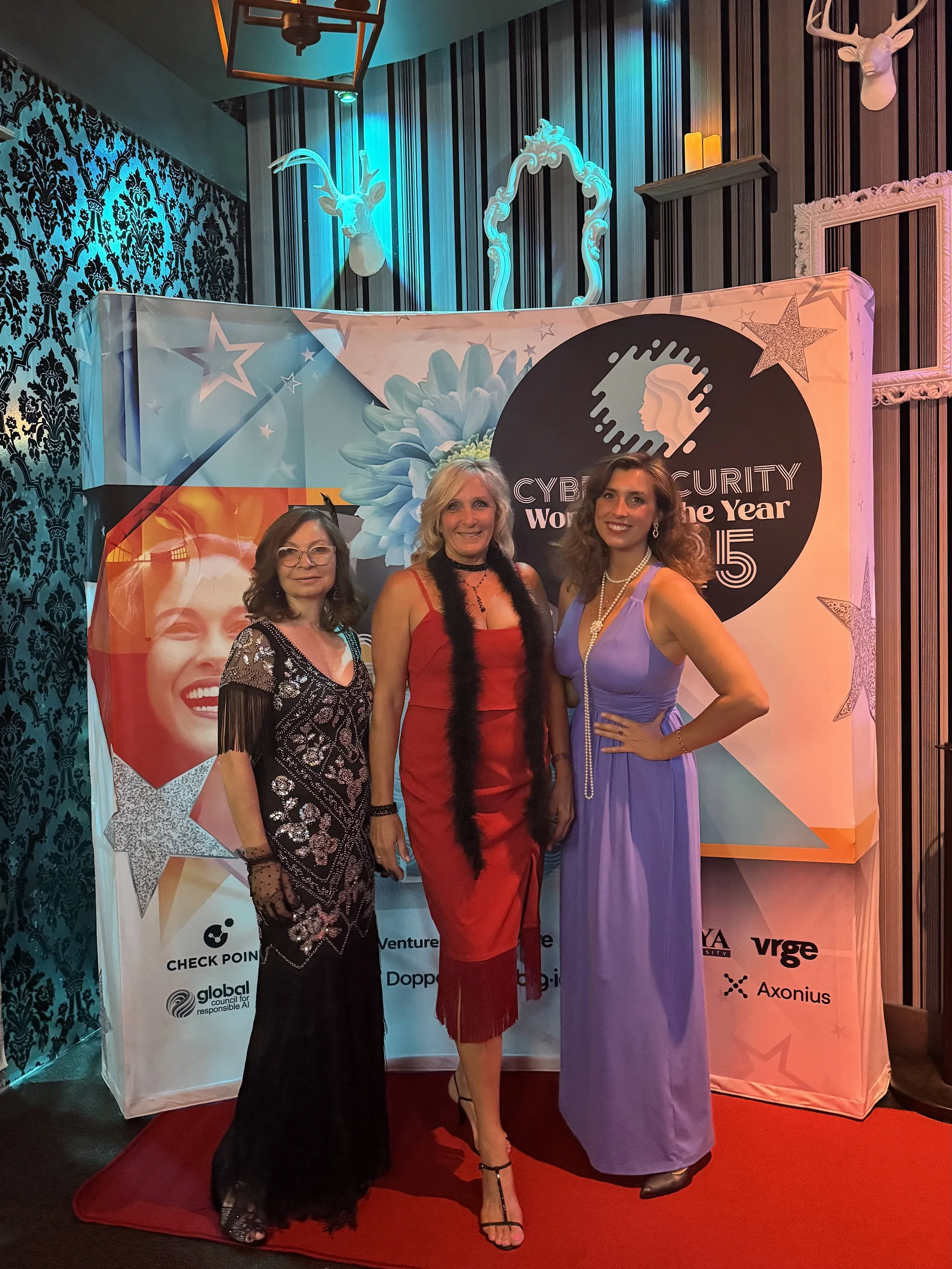 Three women dressed in formal evening attire standing on a red carpet in front of a backdrop at the Cybersecurity Woman of the Year 2015 event. The backdrop features a woman’s face, flowers, and stars.