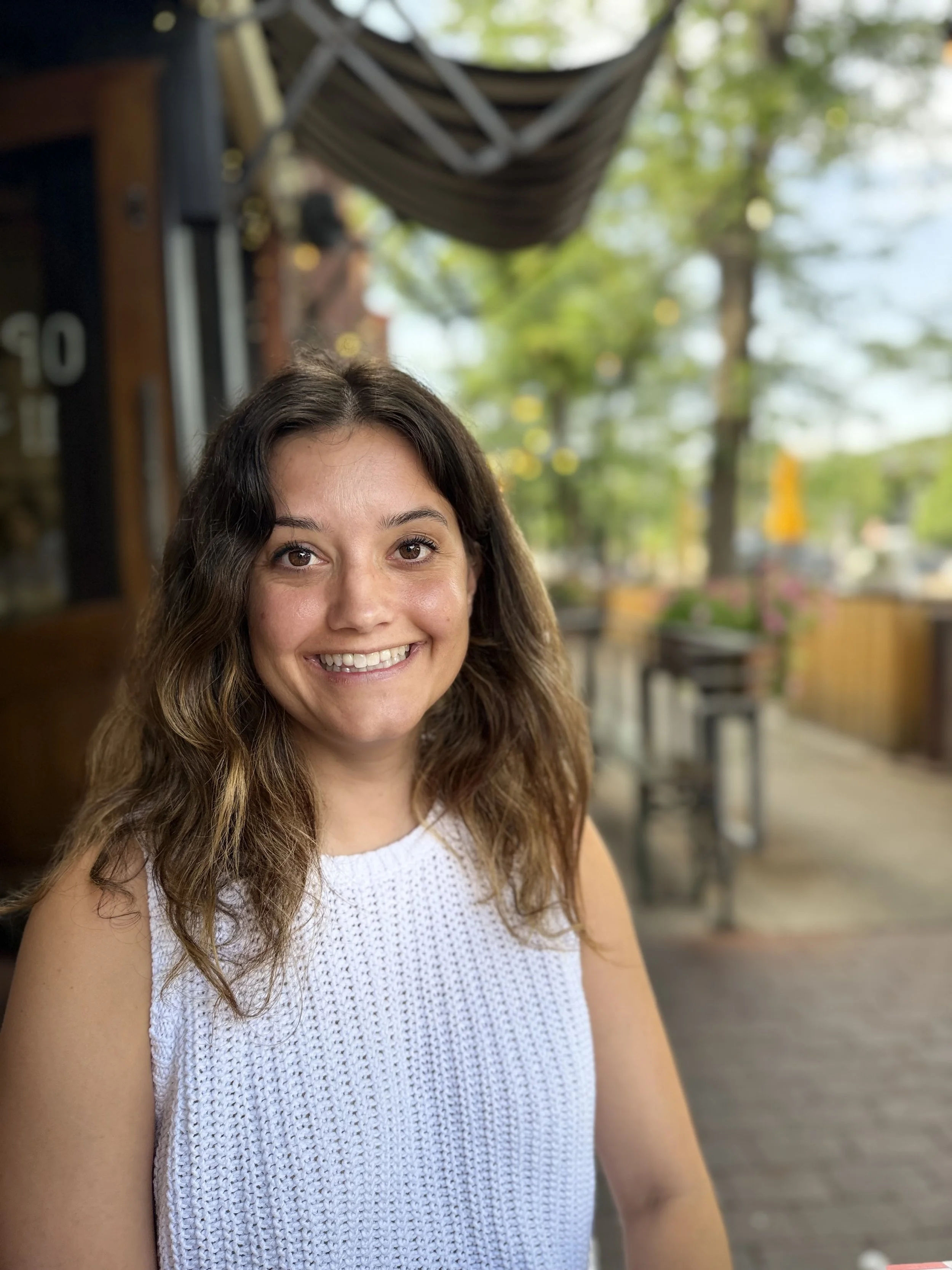 A woman with wavy brown hair and a white sleeveless top smiling on a patio with blurred outdoor furniture and a tree in the background.