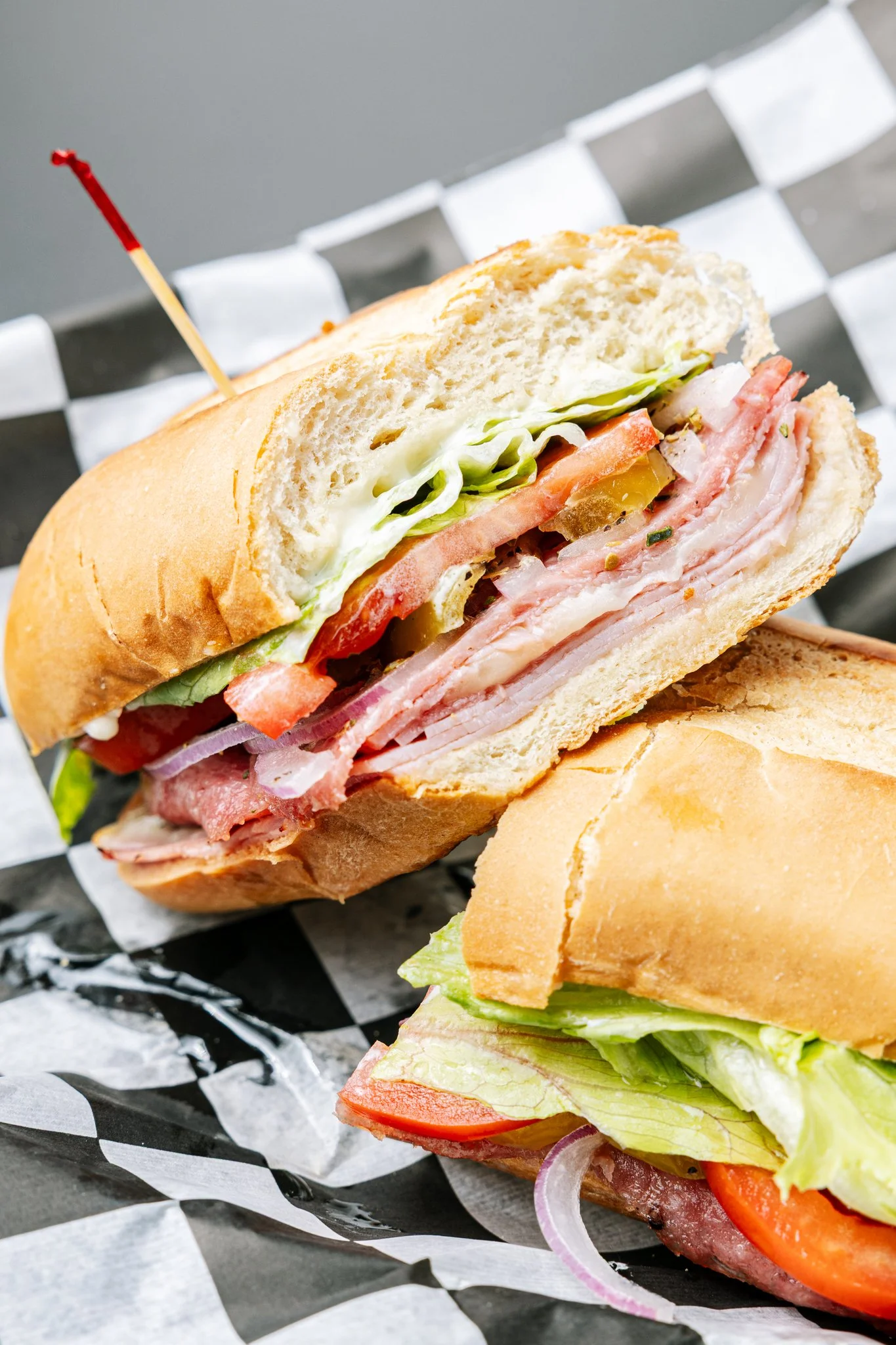 Close-up of a submarine sandwich with deli meats, lettuce, tomato, onion, and pickles inside a hoagie bun, on black and white checkered paper.