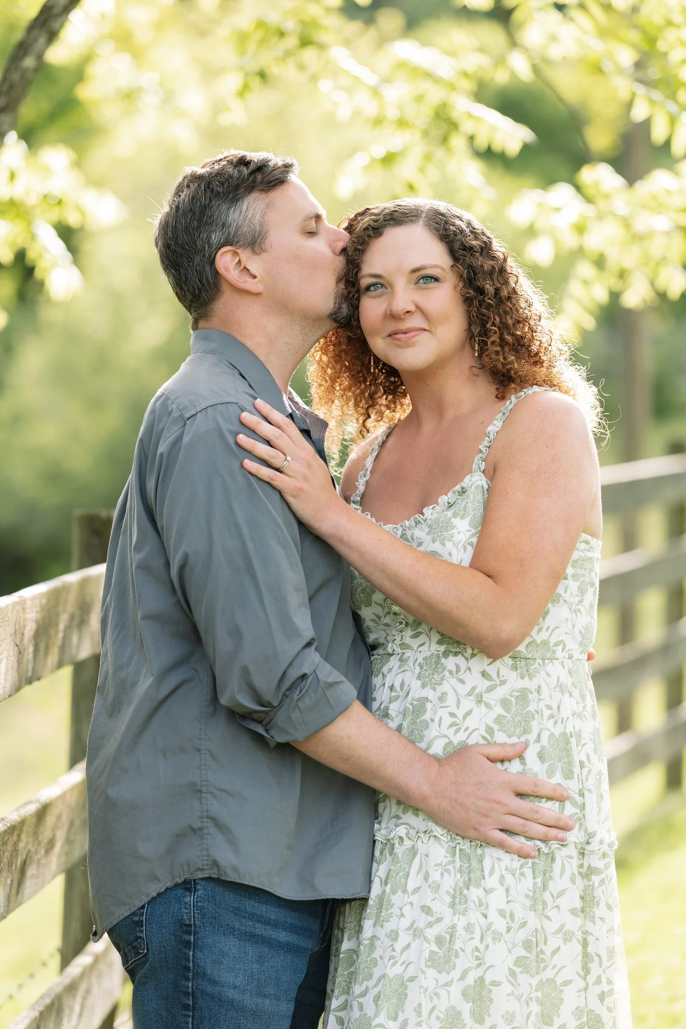 A couple standing outdoors by a wooden fence and overhanging branches together as the male kisses the temple of the female and the female gazes at the camera.