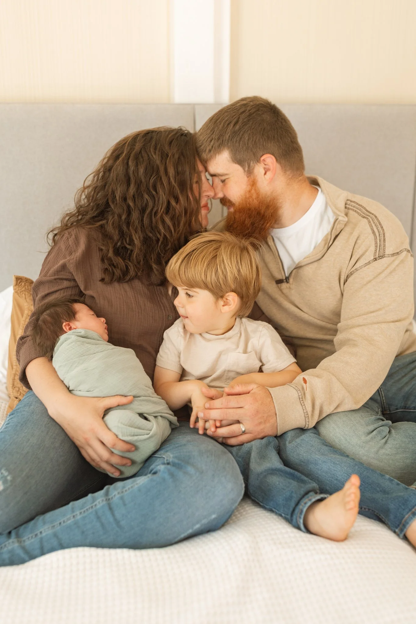 A mom and dad embracing while holding their toddler son and newborn baby boy on their bed during an at-home family session in Tampa, FL.