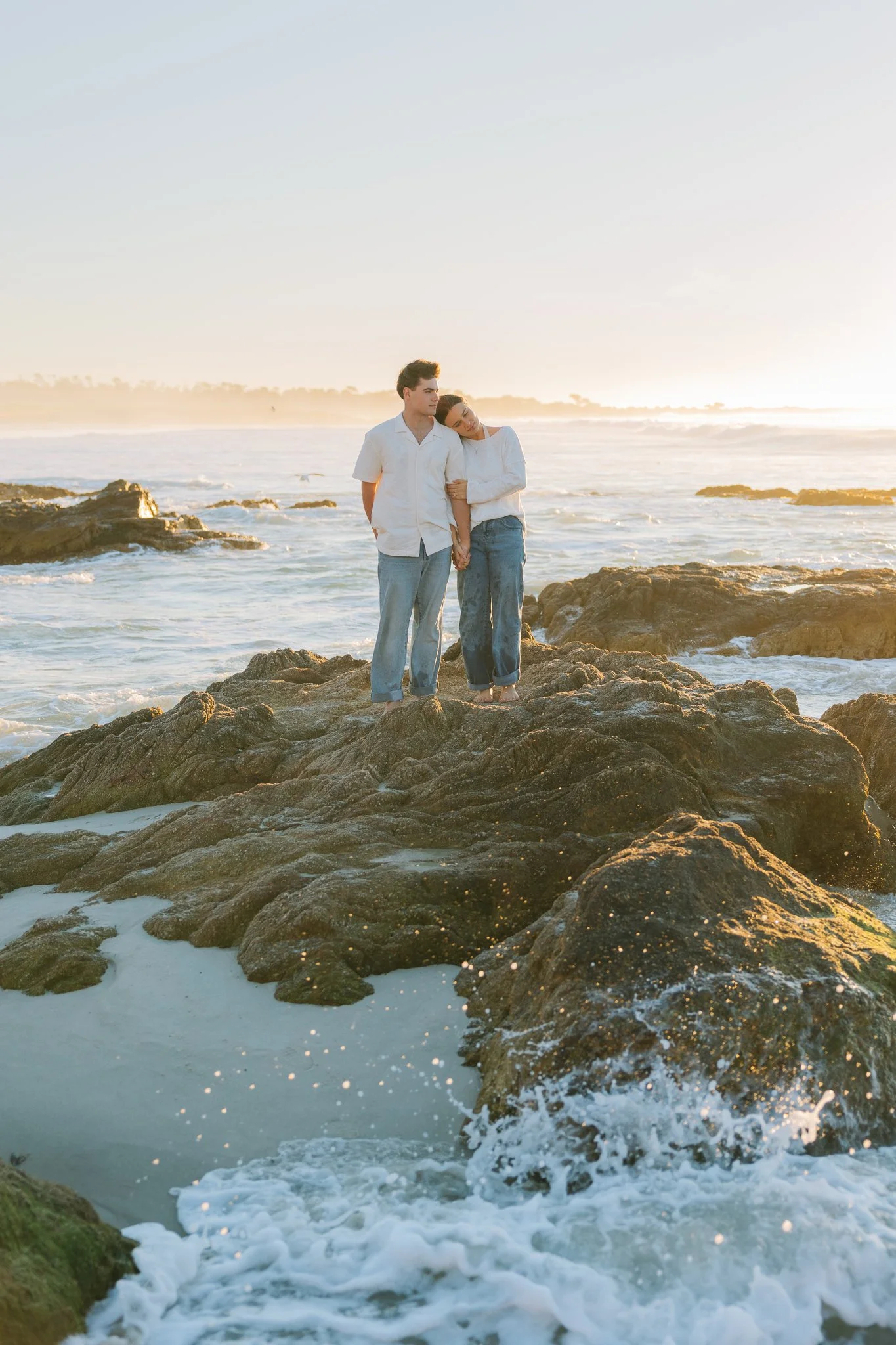 A young couple standing on a rocky shore as the waves splash around them during a Tampa, FL engagement session.