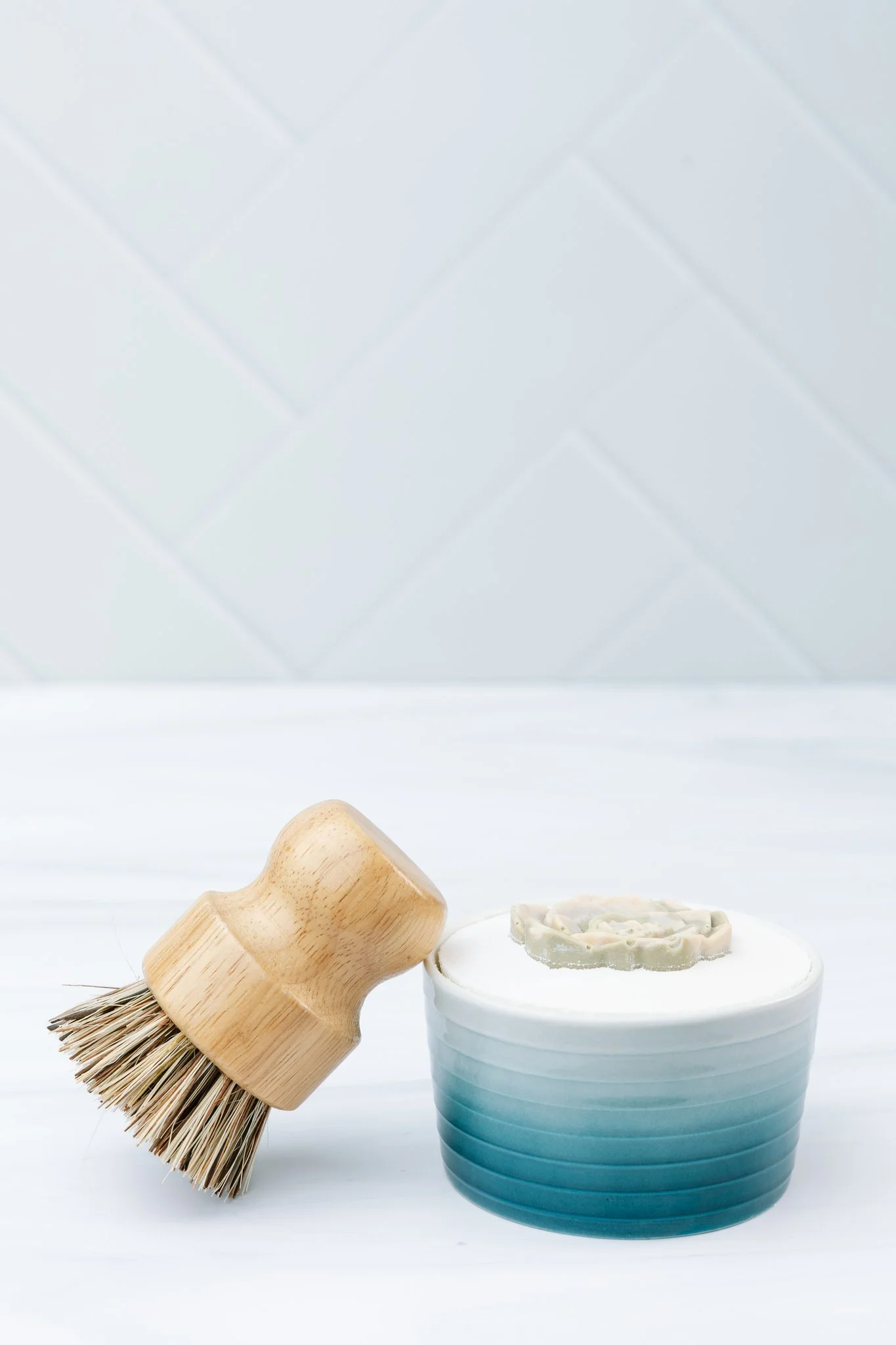 A wooden scrub brush with natural bristles leaning on a white porcelain dish containing a partially visible soap or lotion bar and a white and blue striped ceramic cup