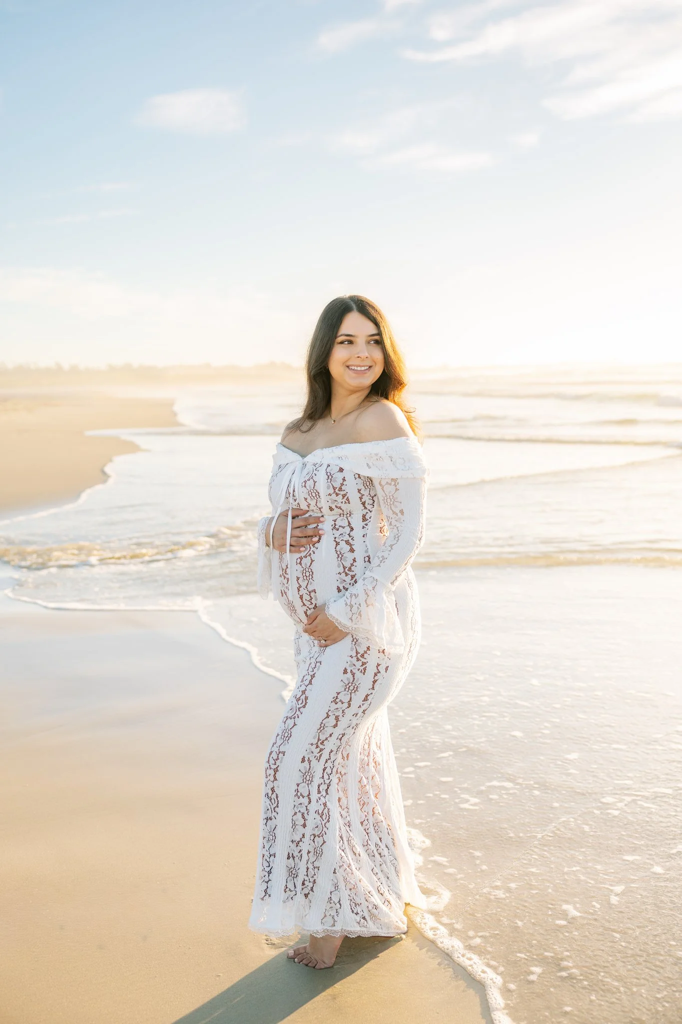 Pregnant woman in white lace maternity dress standing on the beach with waves crashing behind her, caressing her bump, for a maternity session in Apollo Beach, FL.