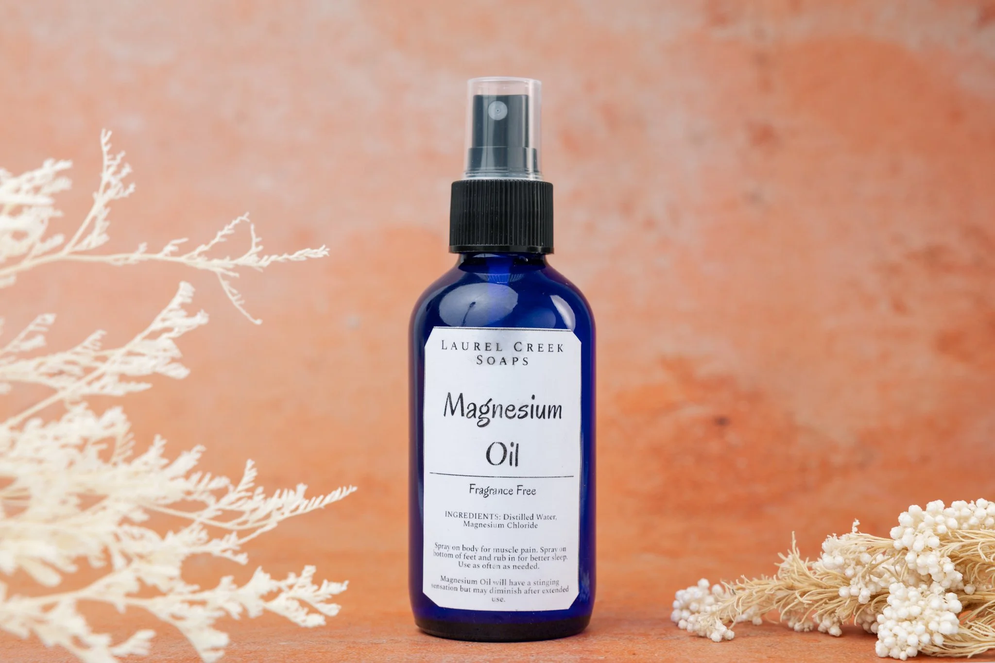 A blue spray bottle labeled 'Magnesium Oil' from Laurel Creek Soaps, standing on a wooden surface with white dried botanical decorations around it, in front of a pinkish background.
