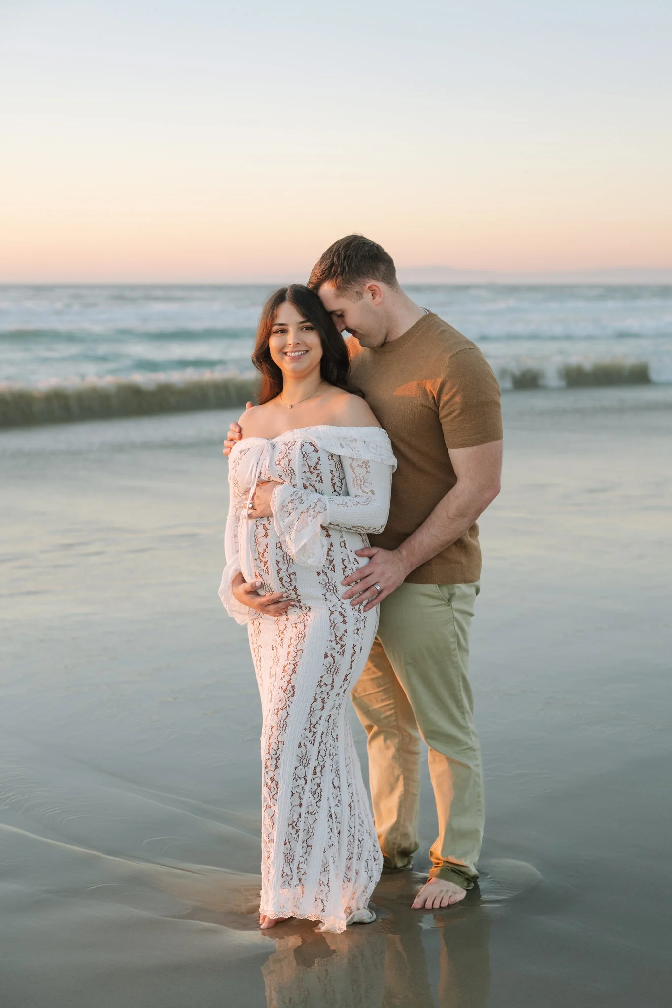 A smiling woman in a white off-the-shoulder lace dress holding a pregnant belly, and a man in a brown t-shirt and khaki pants standing behind her on the beach at sunset, with the ocean in the background during a Tampa, FL maternity session.