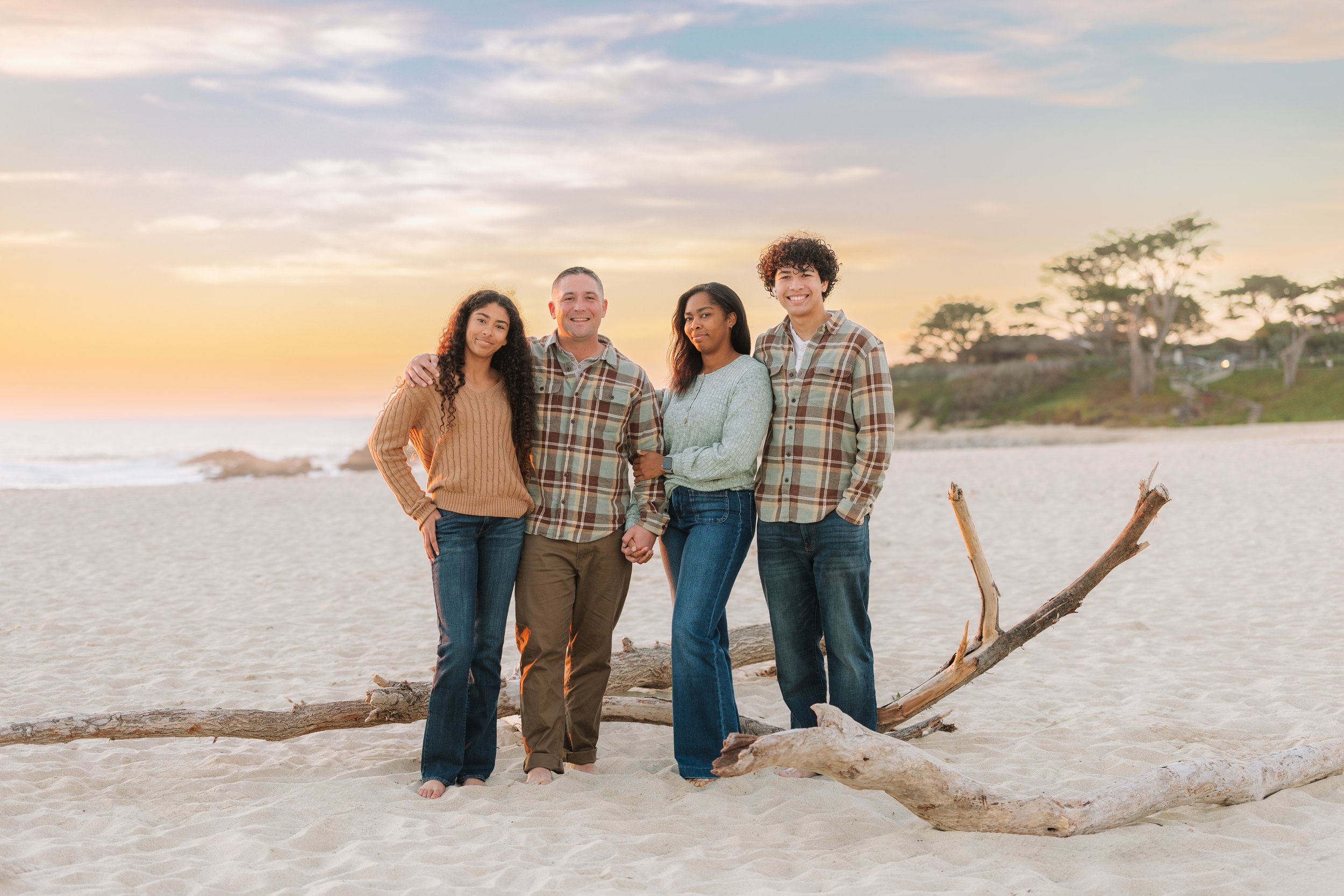 A family of 4, mother, father, and 2 teenage children, standing on the sandy beach by driftwood, smiling at the camera.