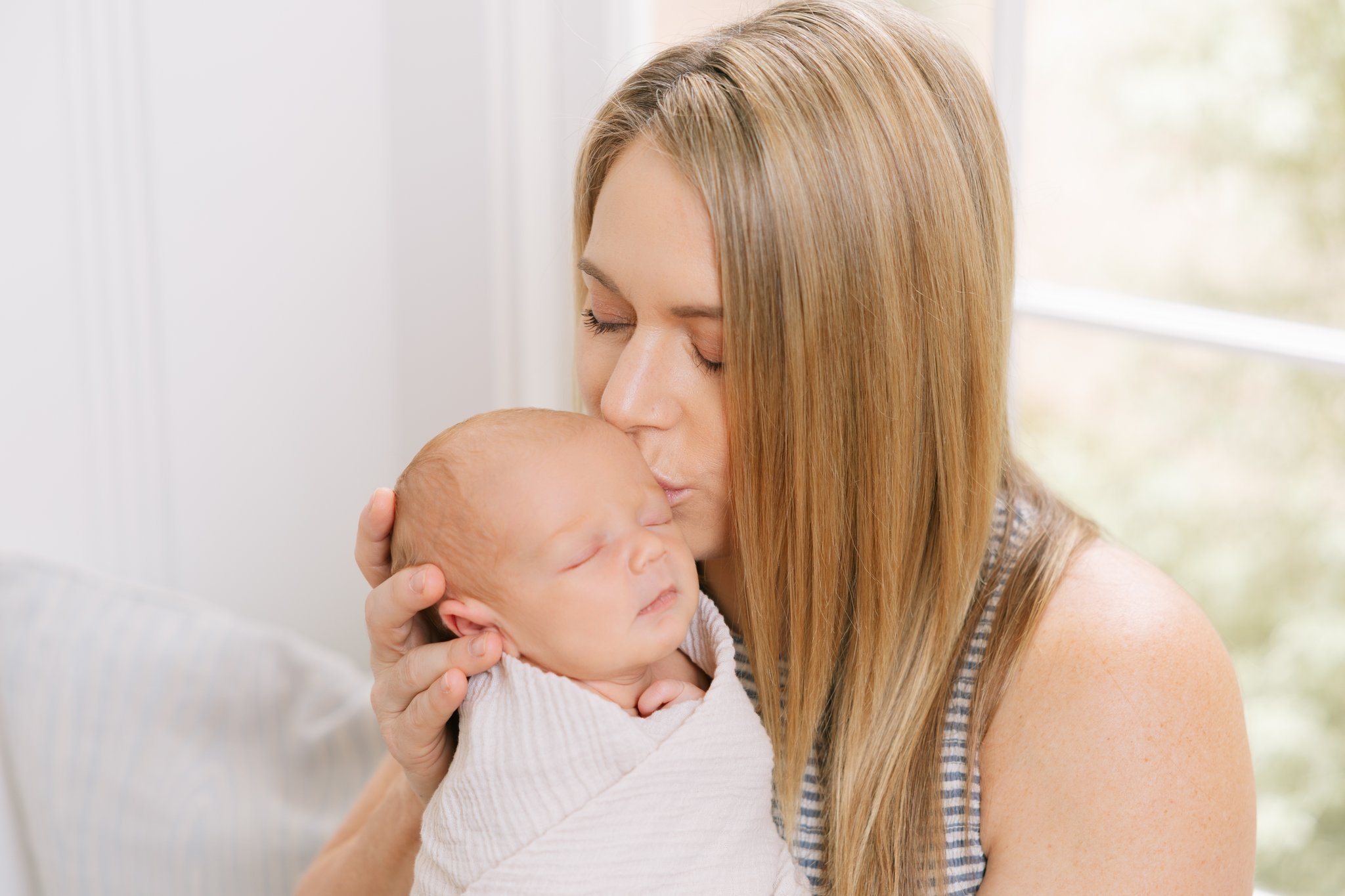 Close up of mother kissing the head of her newborn baby, swaddled in a cream wrap, during an at-home newborn session in Tampa, FL.
