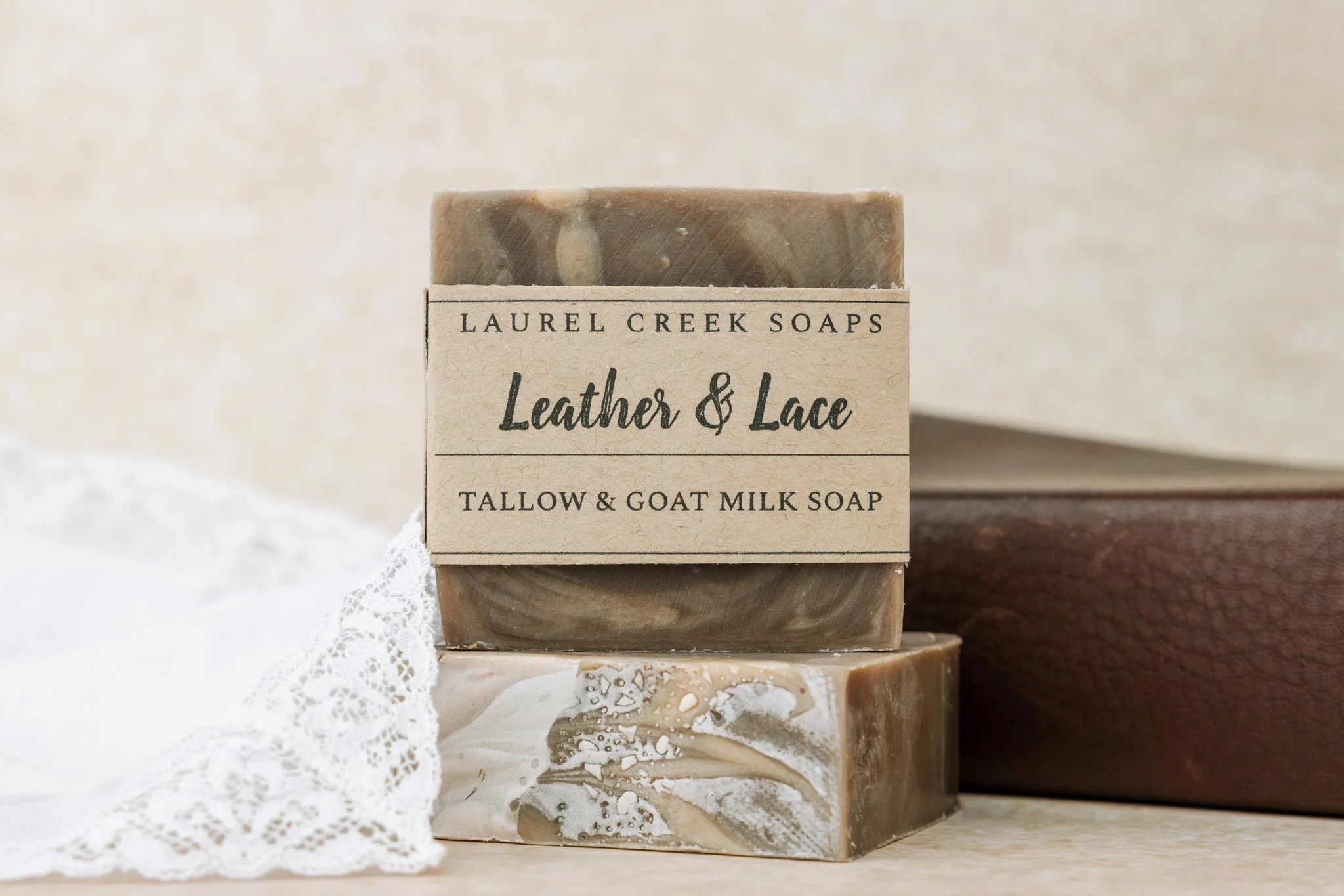 Bar of soap labeled 'Laurel Creek Soaps Leather & Lace Tallow & Goat Milk Soap' placed on a lace cloth with a neutral background.