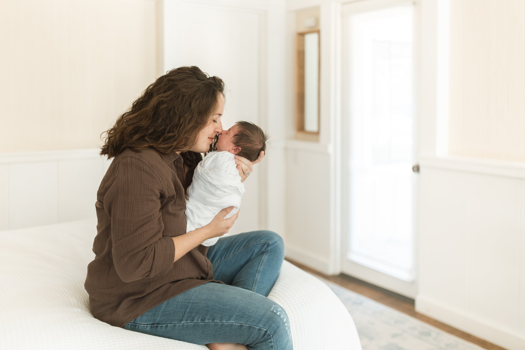A mother embracing her newborn son, sitting on the edge of her bed, nose-to-nose with baby during an at-home newborn session in Tampa, FL.