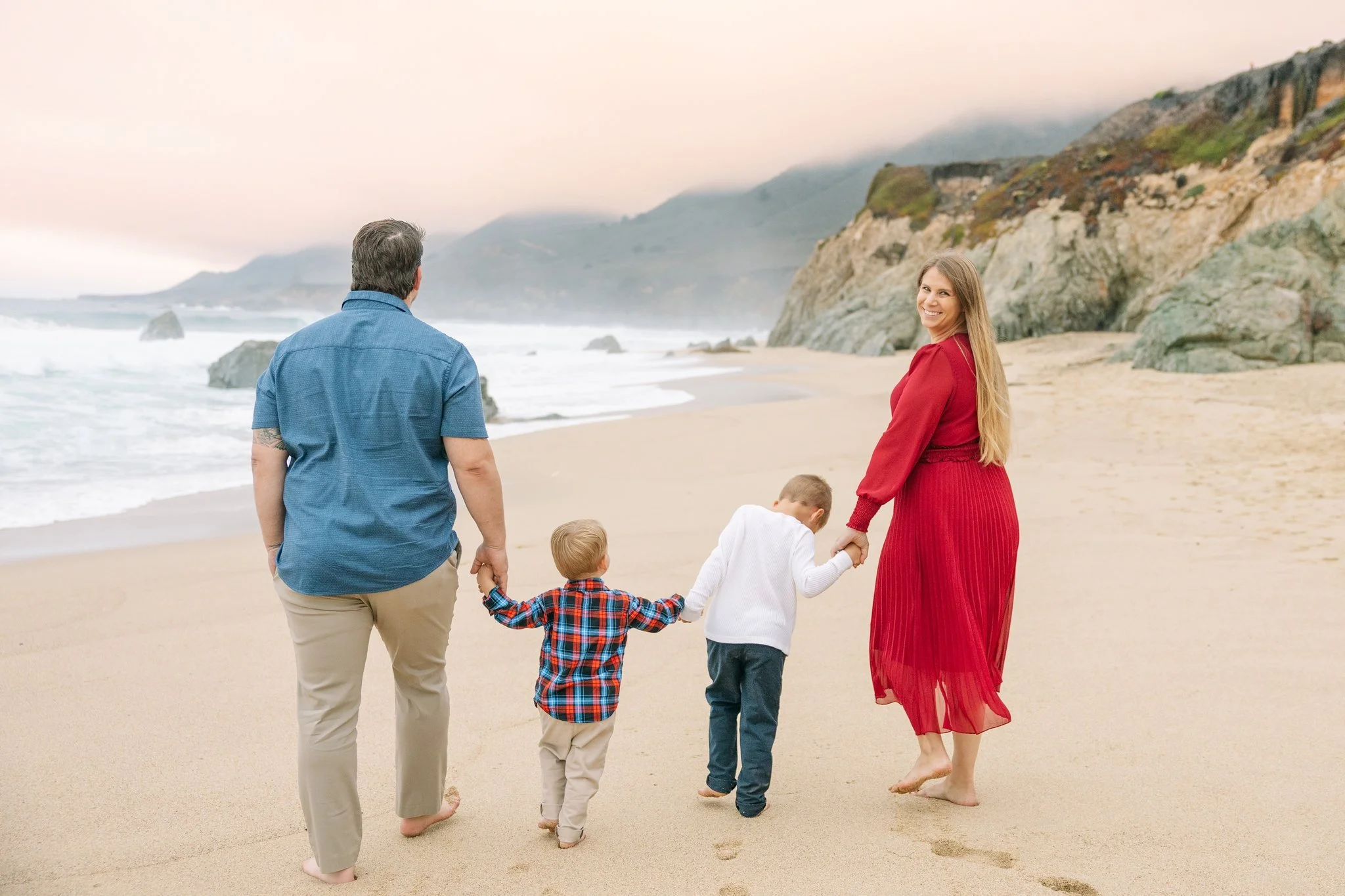 Family of 4 walking on a beach at sunset away from the camera, while the mom looks over her shoulder back at the camera, smiling.