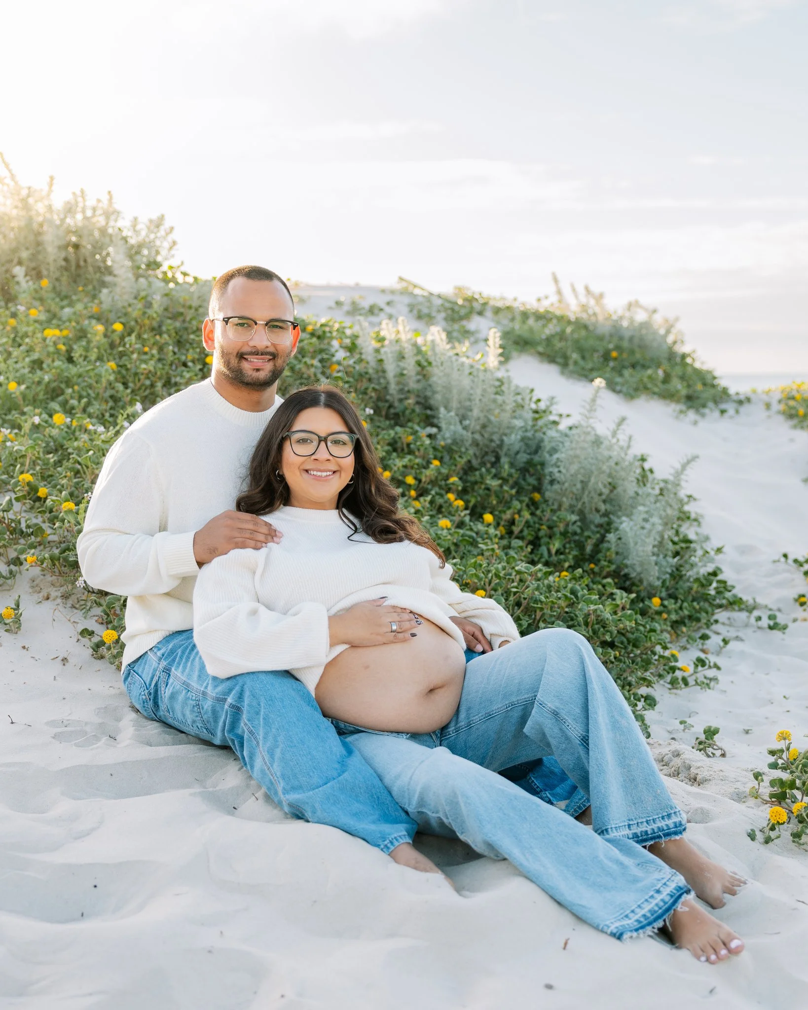Couple sitting in the sand together, smiling at the camera, posing for beach maternity photos in Tampa, FL .