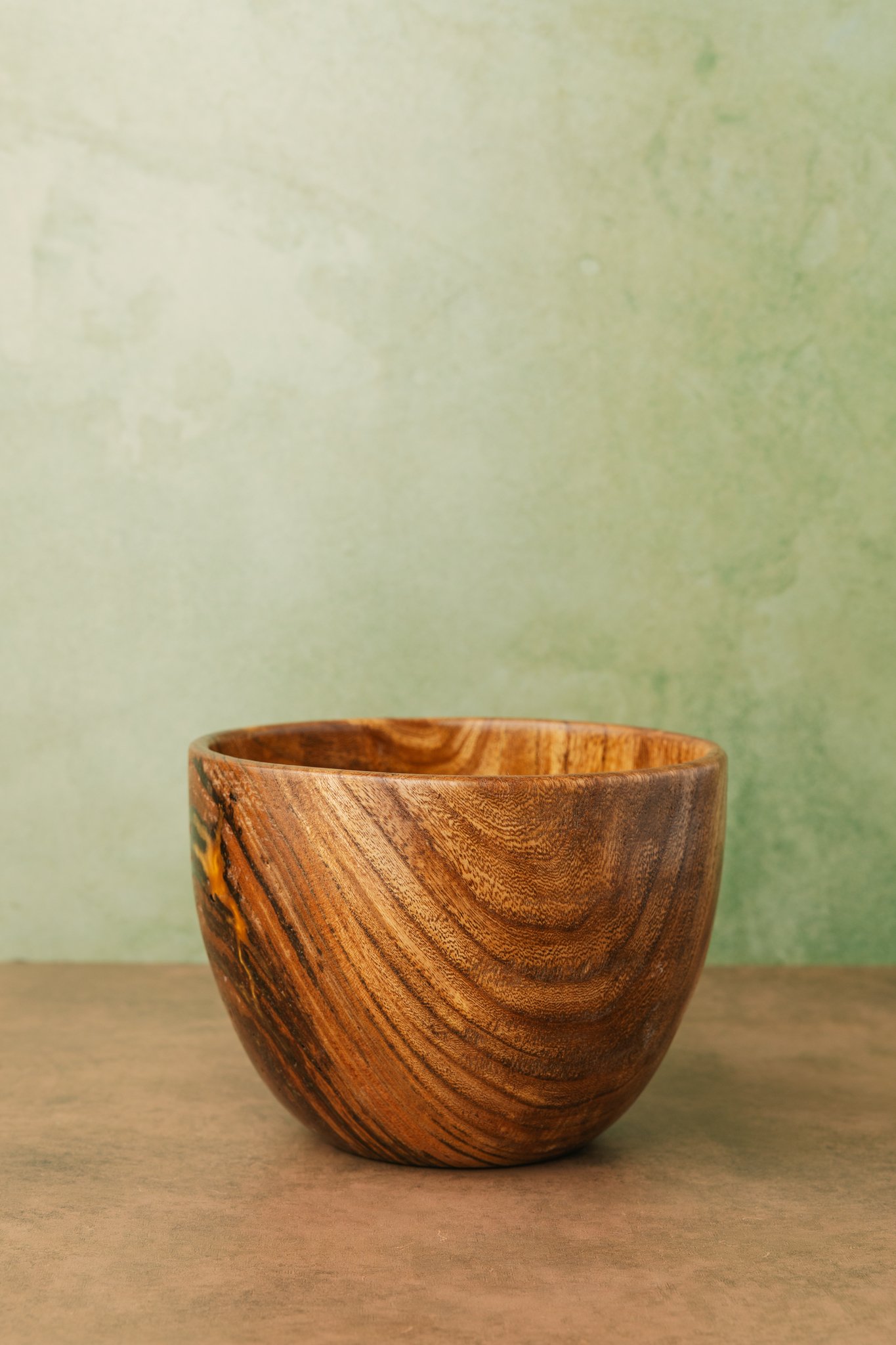 A smooth, polished wooden bowl with a natural wood grain pattern, placed on a wooden surface against a pale, textured green background.