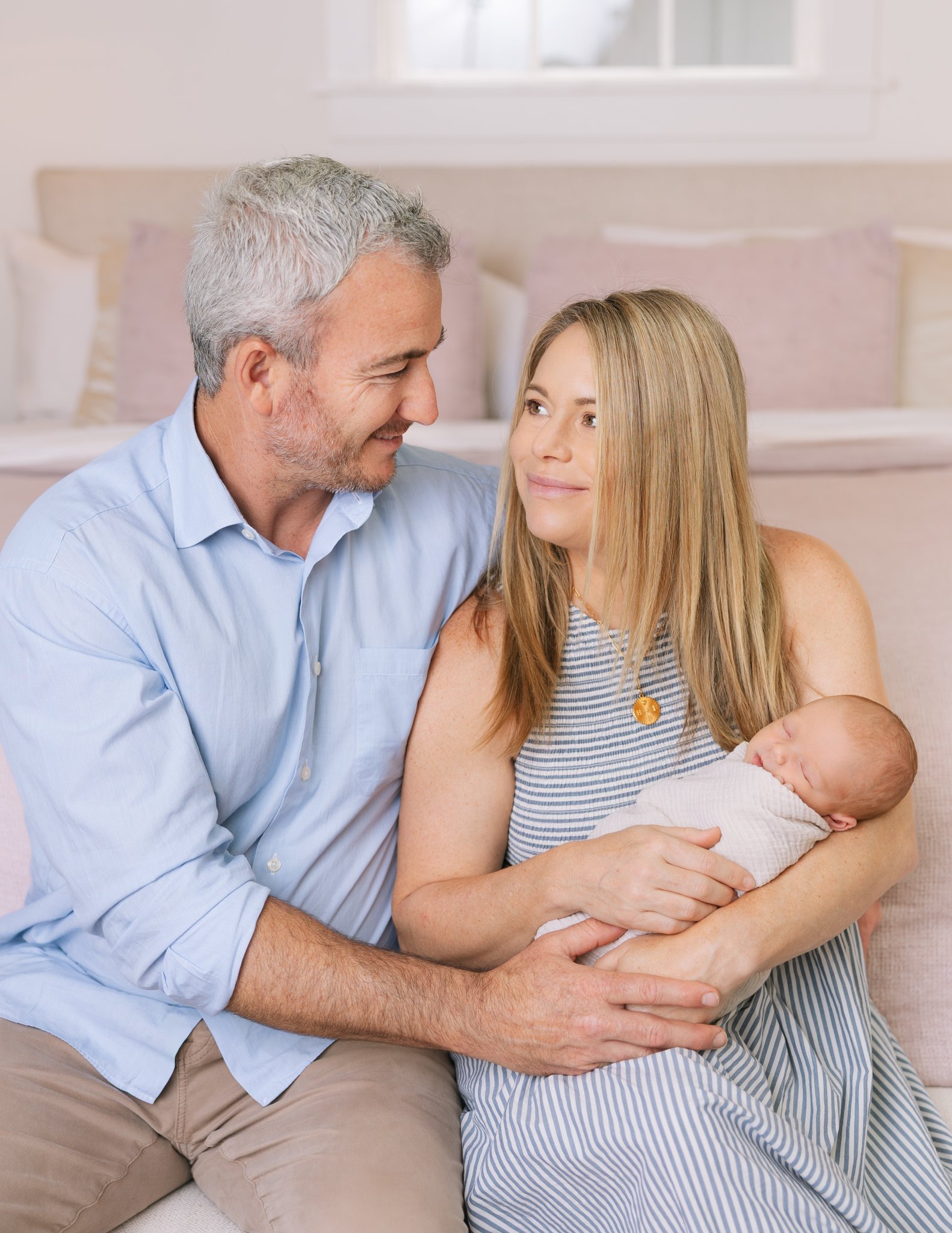 Father and mother smiling at each other, holding newborn baby girl, sitting on their bed, during their at-home newborn session in Tampa, FL.