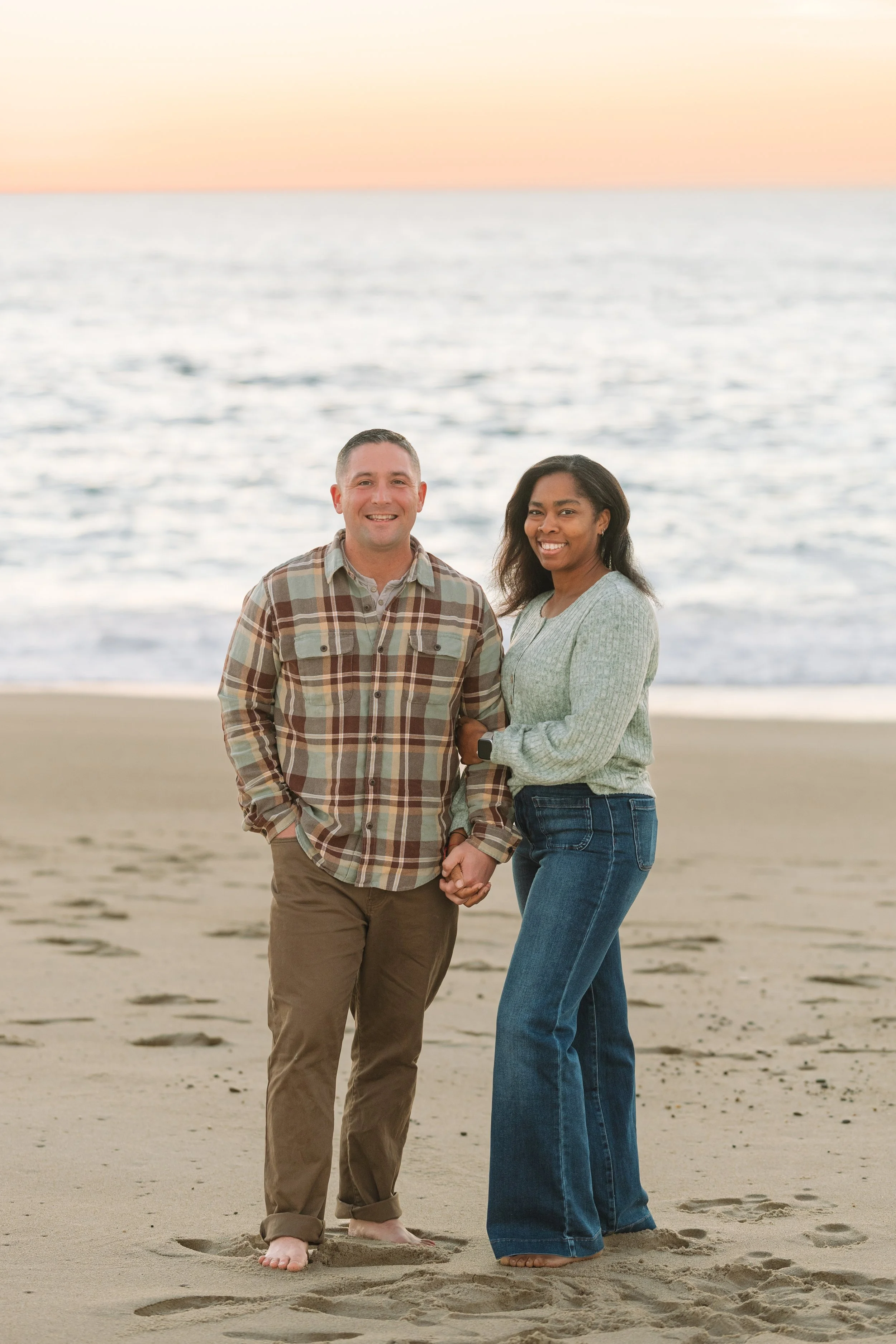 A smiling man and woman holding hands on the beach during sunset, standing in shallow water with footprints in the sand.