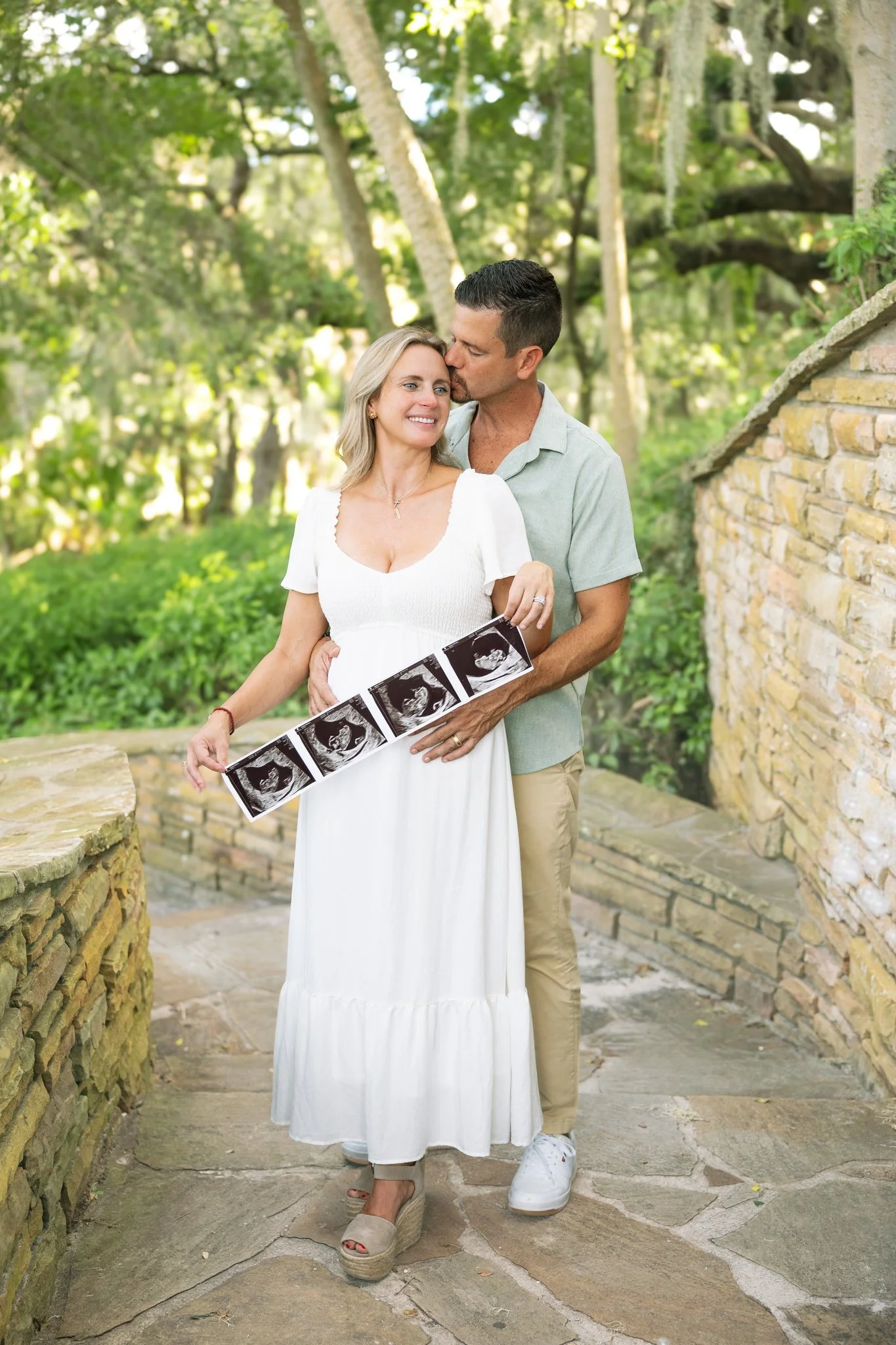 Husband kissing his wife's temple as she holds an ultrasound strip along a stone-walled path at Philippe Park