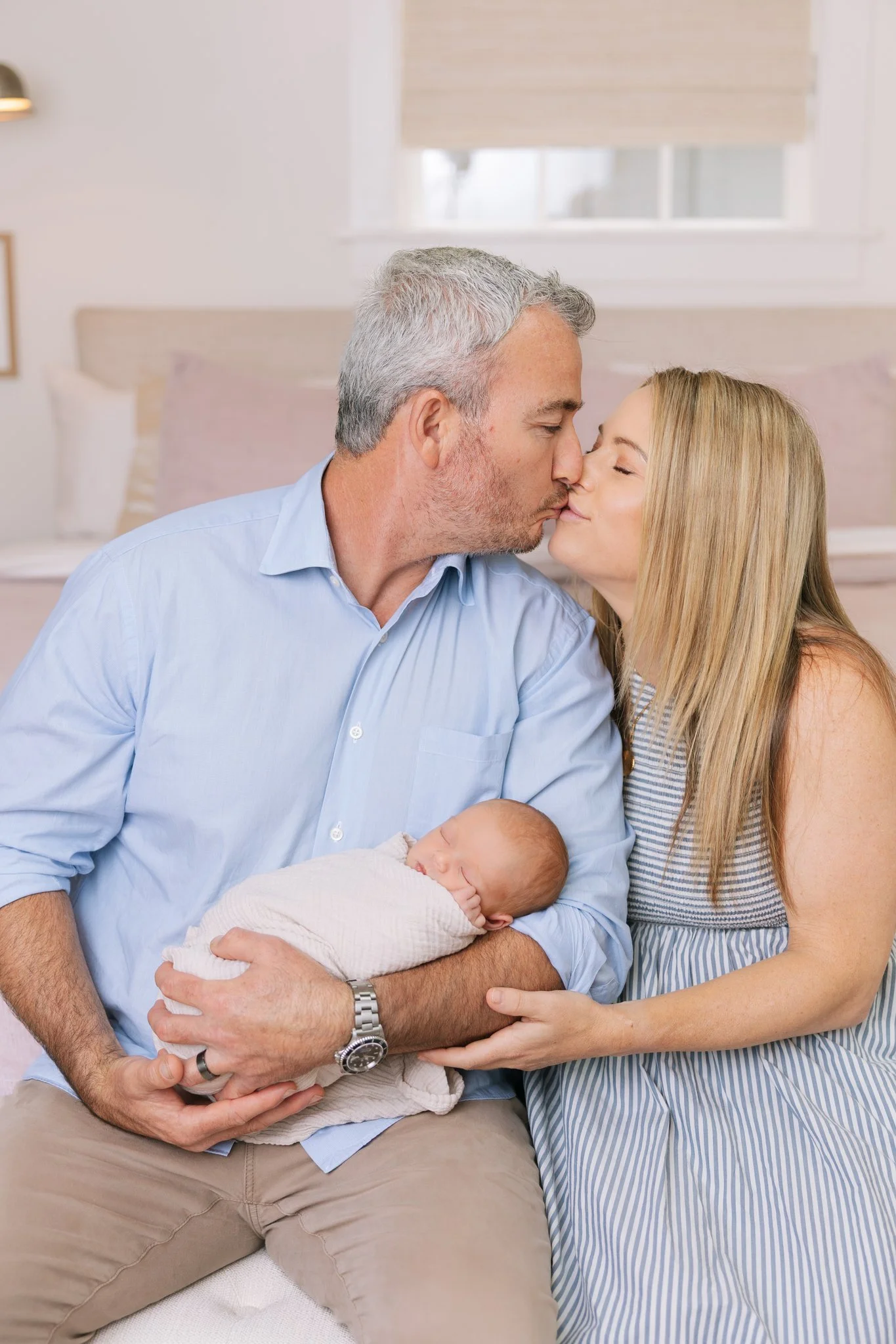 Father and mother kissing each other while holding newborn baby girl, sitting on their bed, during their at-home newborn session in Tampa, FL.