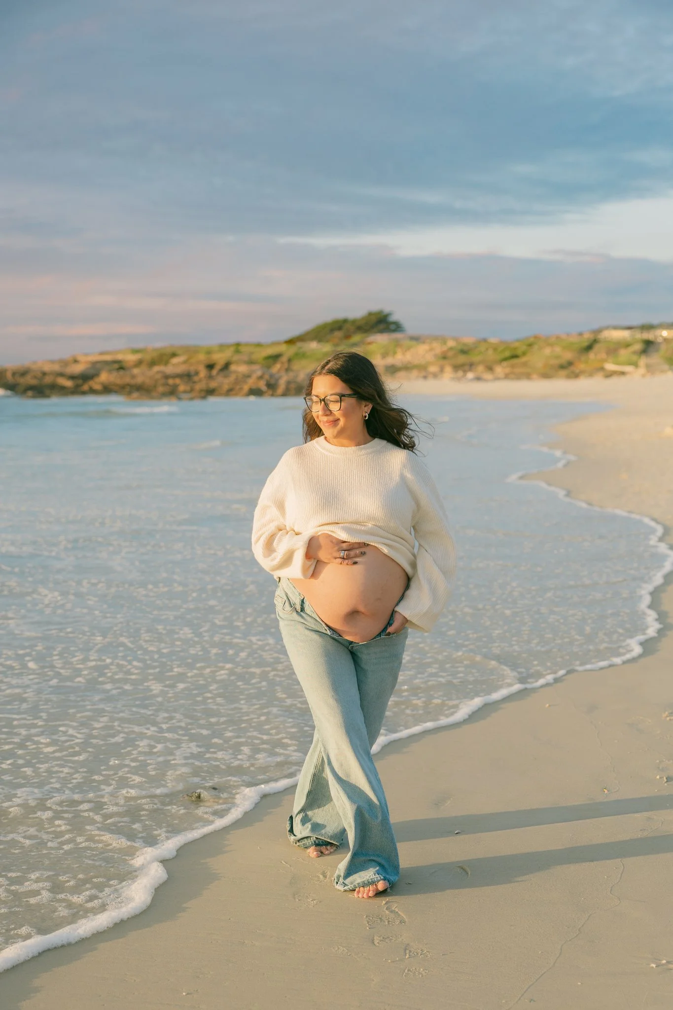 Pregnant woman walking on the beach by the waves, smiling at the water for her beach maternity photos in Tampa, FL.