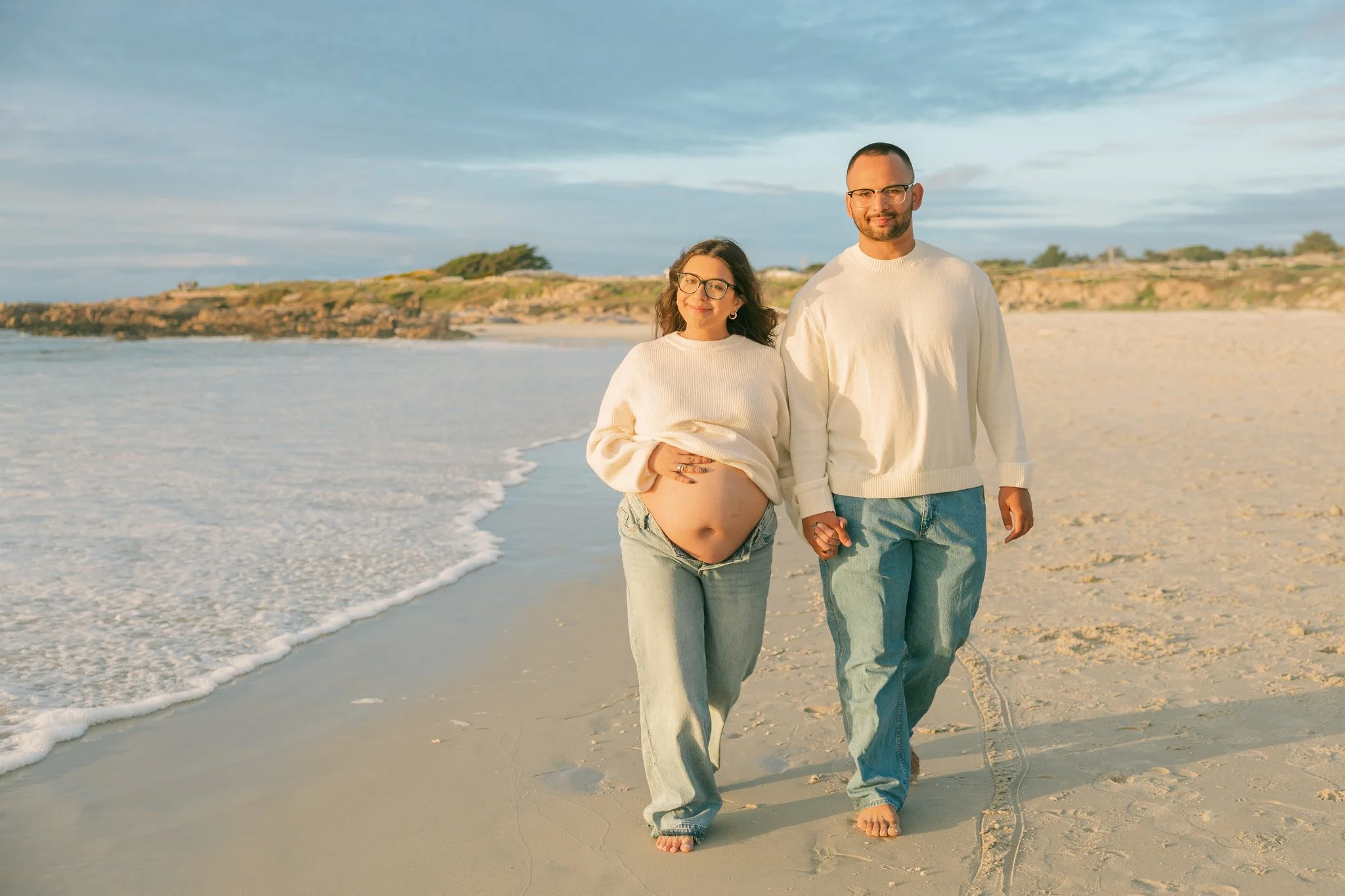 A couple walking on the beach, holding hands and smiling at the camera as a wave comes rushing in beside them.