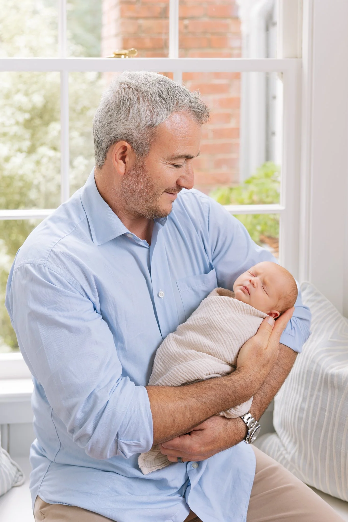 Father holding his newborn baby, sitting at a window bench in their home during an at-home newborn session in Tampa, FL.