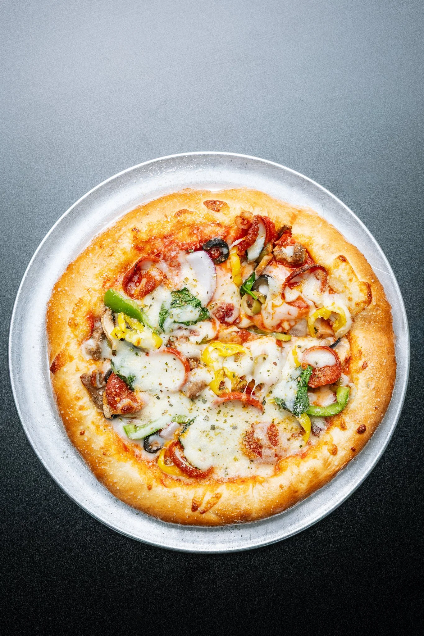 Partial view of a pepperoni pizza with colorful toppings on a silver tray, placed on a dark surface.