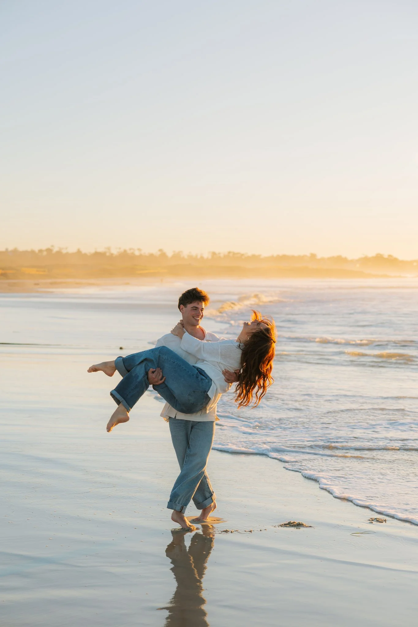 Couple on the beach at sunset being playful and smiling as the man picks up the woman and carries her on the beach.