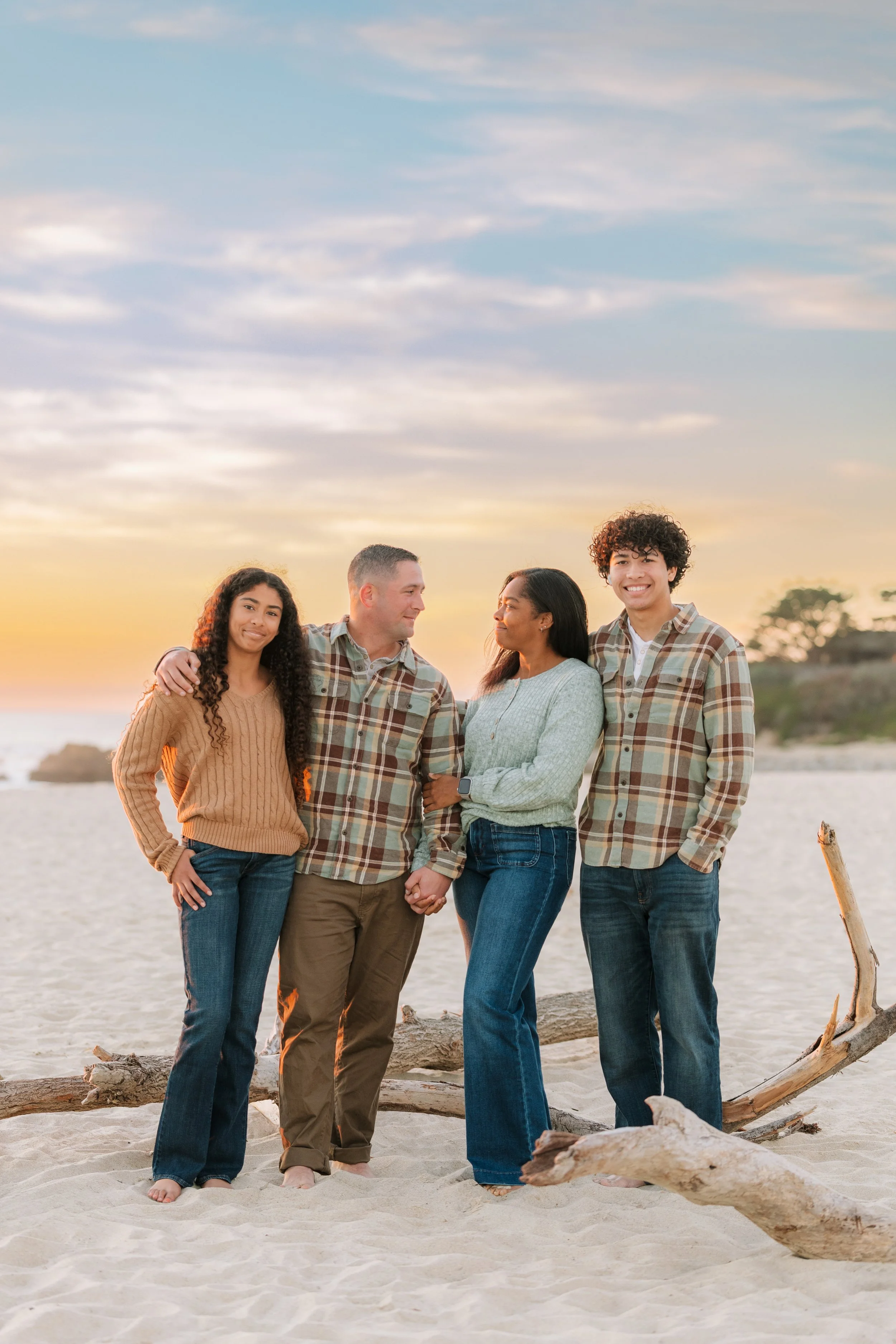 Family of 4 with teenage daughter, father, mother, and teenage son standing on the beach in front of driftwood at sunset for a family session in Apollo Beach, FL.