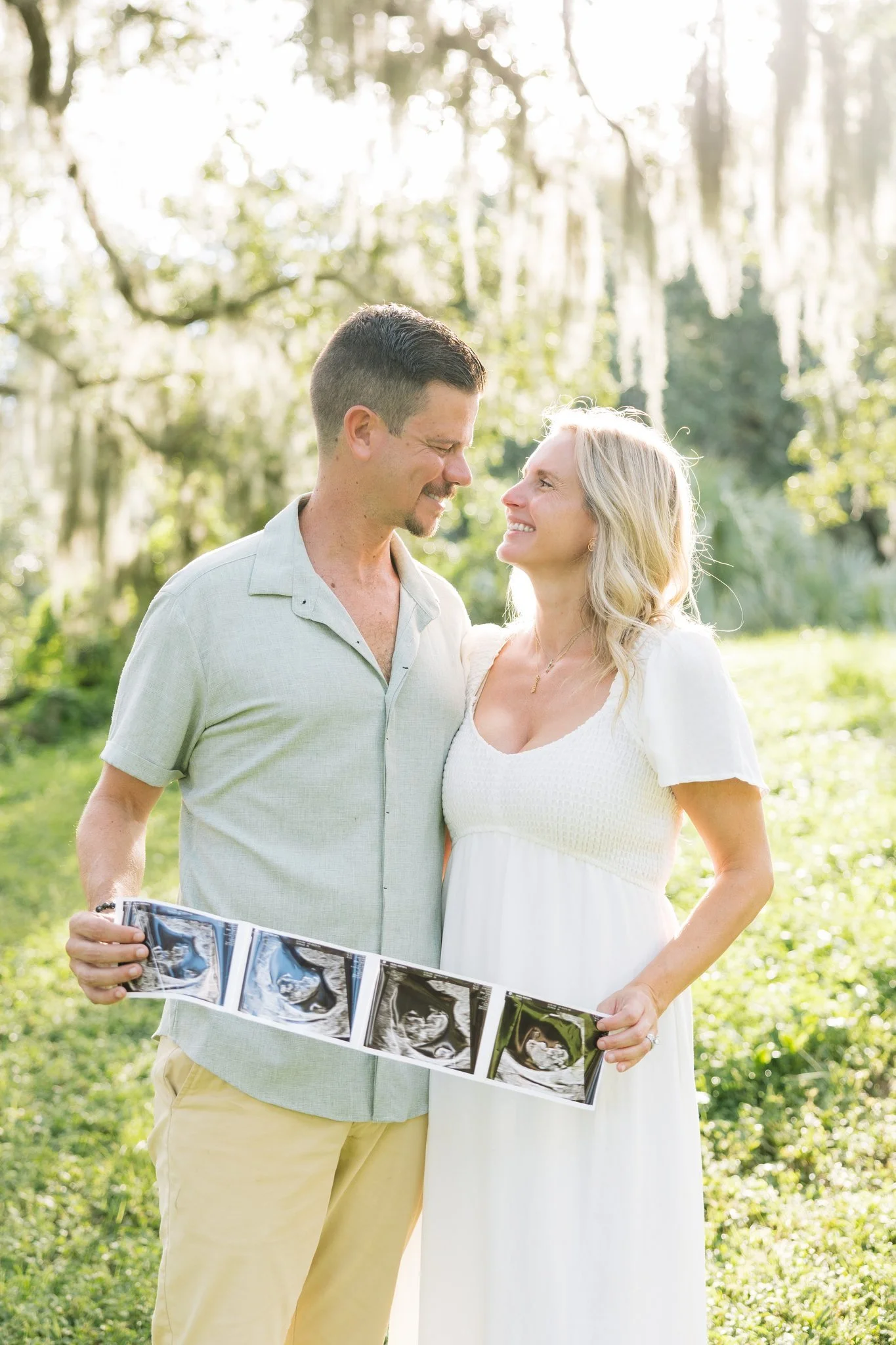 Expecting couple smiling at each other while holding an ultrasound strip under the oaks at Philippe Park in Safety Harbor